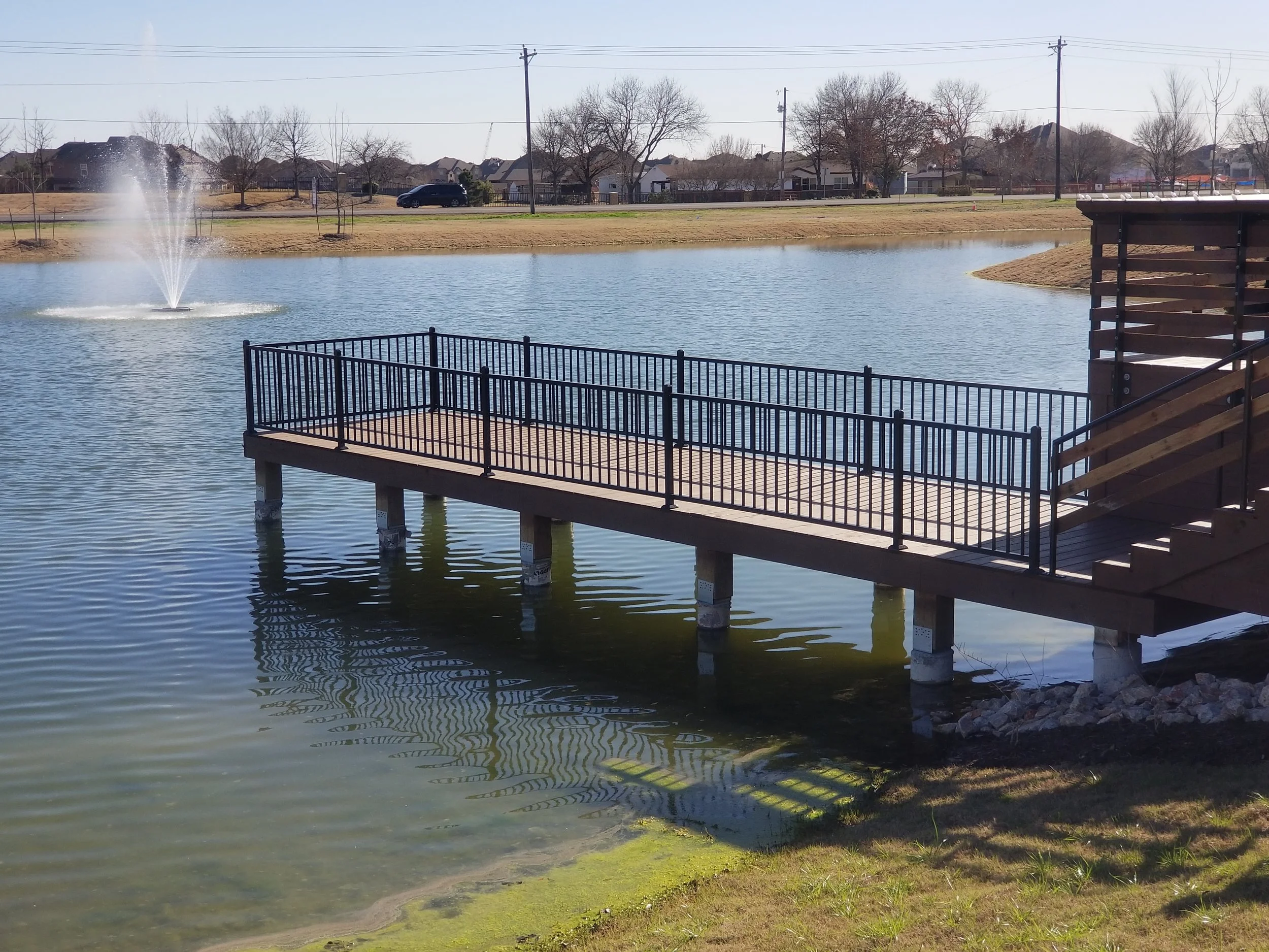 A wooden dock with metal railings extends over a small pond. A fountain sprays water into the air in the pond, and trees and houses are visible in the background under a clear sky.