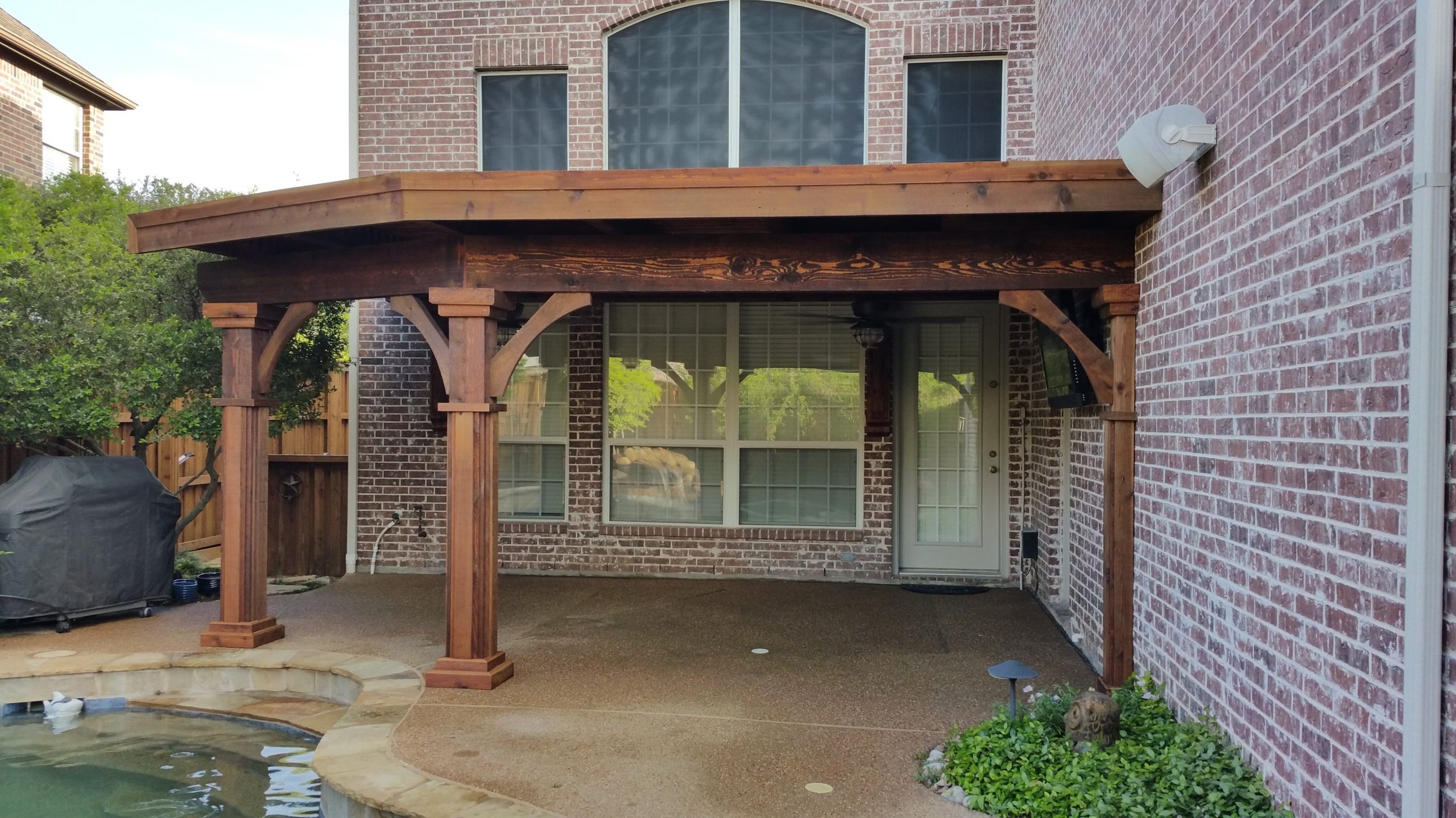 Backyard patio with a covered wooden pergola structure attached to a brick house. The patio has a textured concrete floor and a small garden with plants and a stone frog sculpture near the house wall. A grill covered with a black cover is on the left