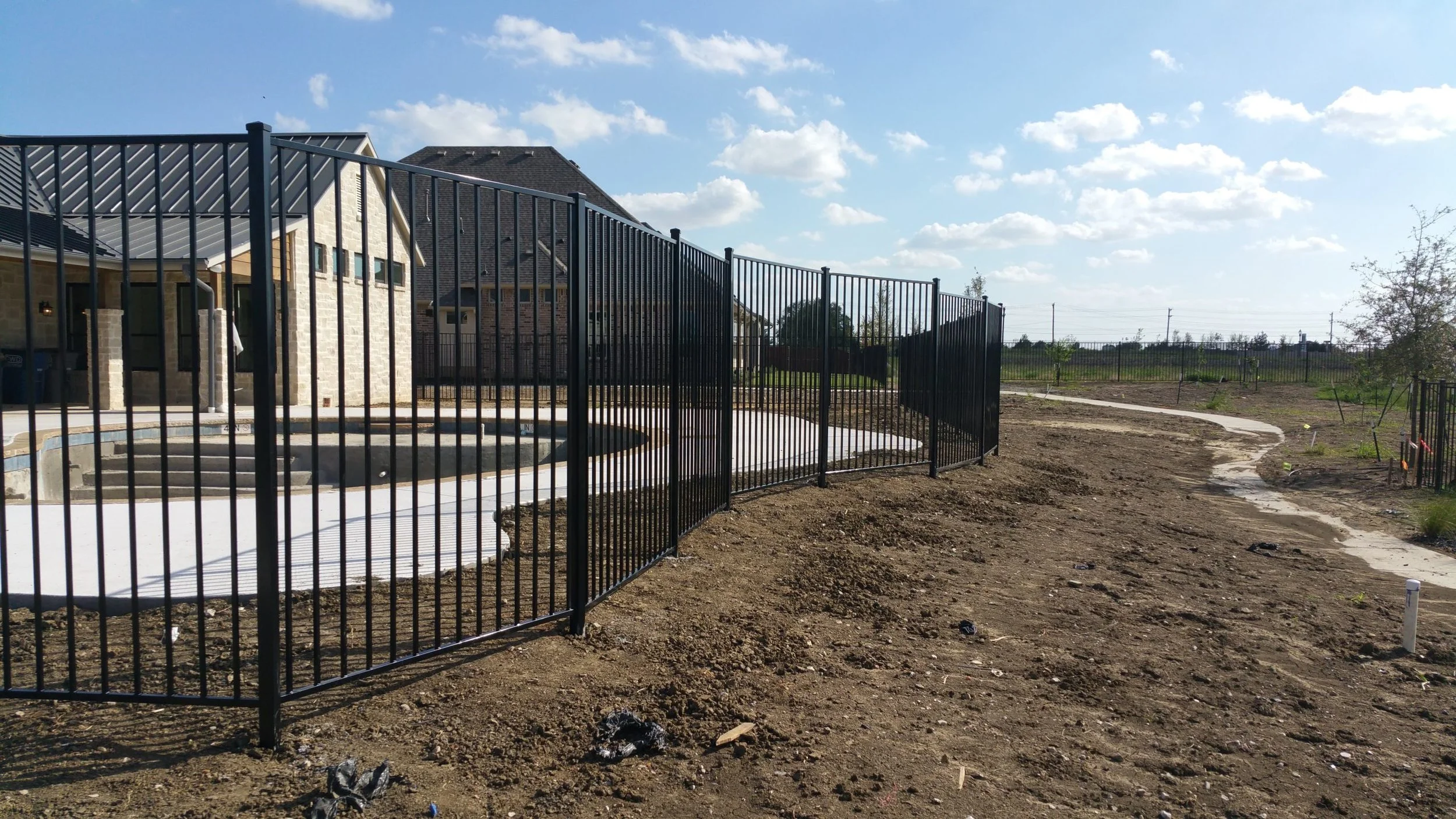 A black metal fence surrounds a backyard pool area, with a concrete pathway leading away from it. The backyard is still under landscaping, with exposed soil and a few small plants, and houses are visible in the background under a partly cloudy sky.