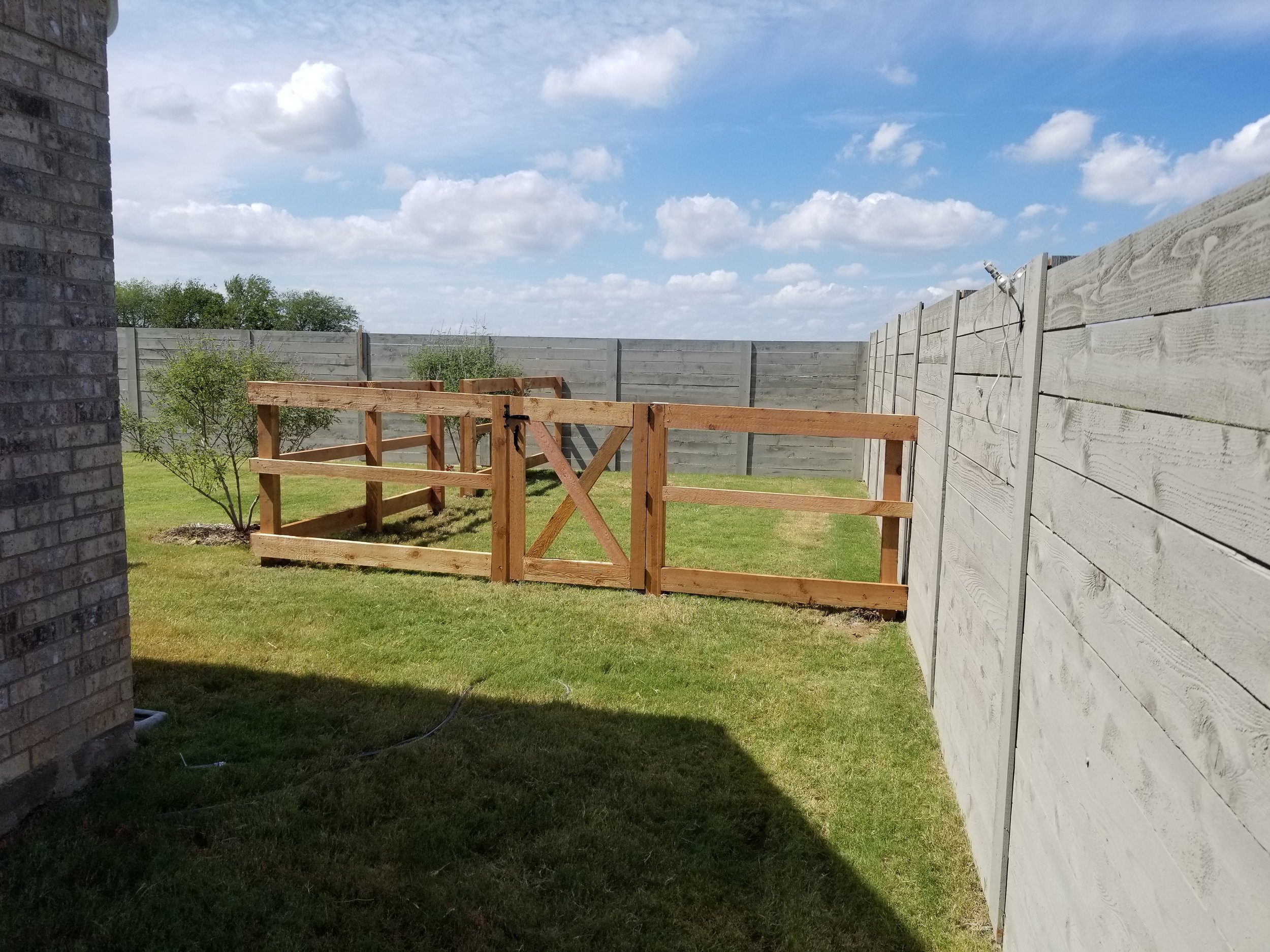 A backyard with new wooden fence panels and a gate, surrounded by a grassy lawn and a gray wooden privacy fence on three sides, under a partly cloudy sky.