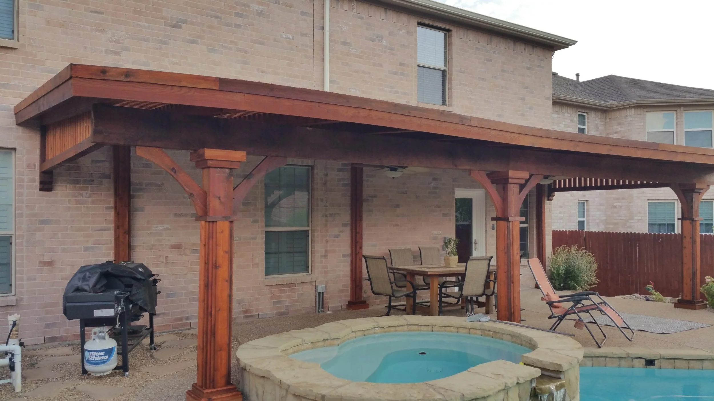 Backyard area with covered patio, outdoor dining table with five chairs, lounge chair, small hot tub, grill, and garden plants, enclosed by a wooden fence, brick house in the background.