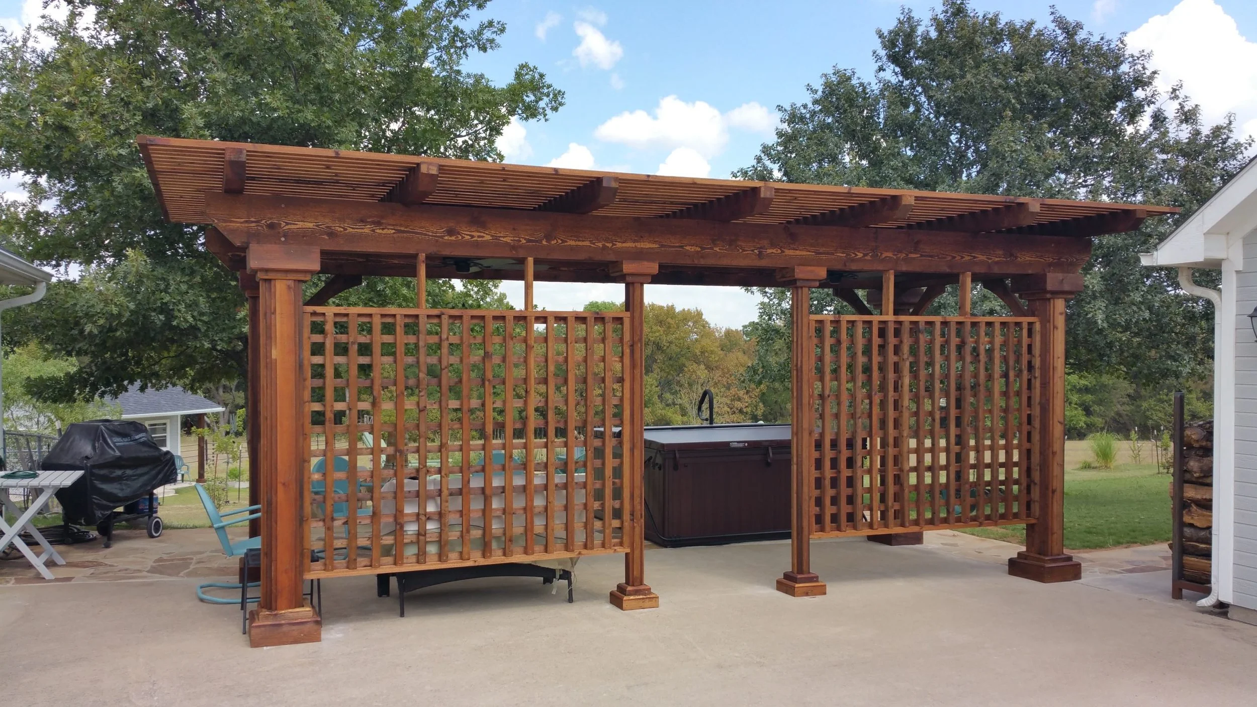 A wooden outdoor gazebo with lattice sides, a slatted roof, and a hot tub inside. There are trees, a grill, and outdoor furniture nearby.