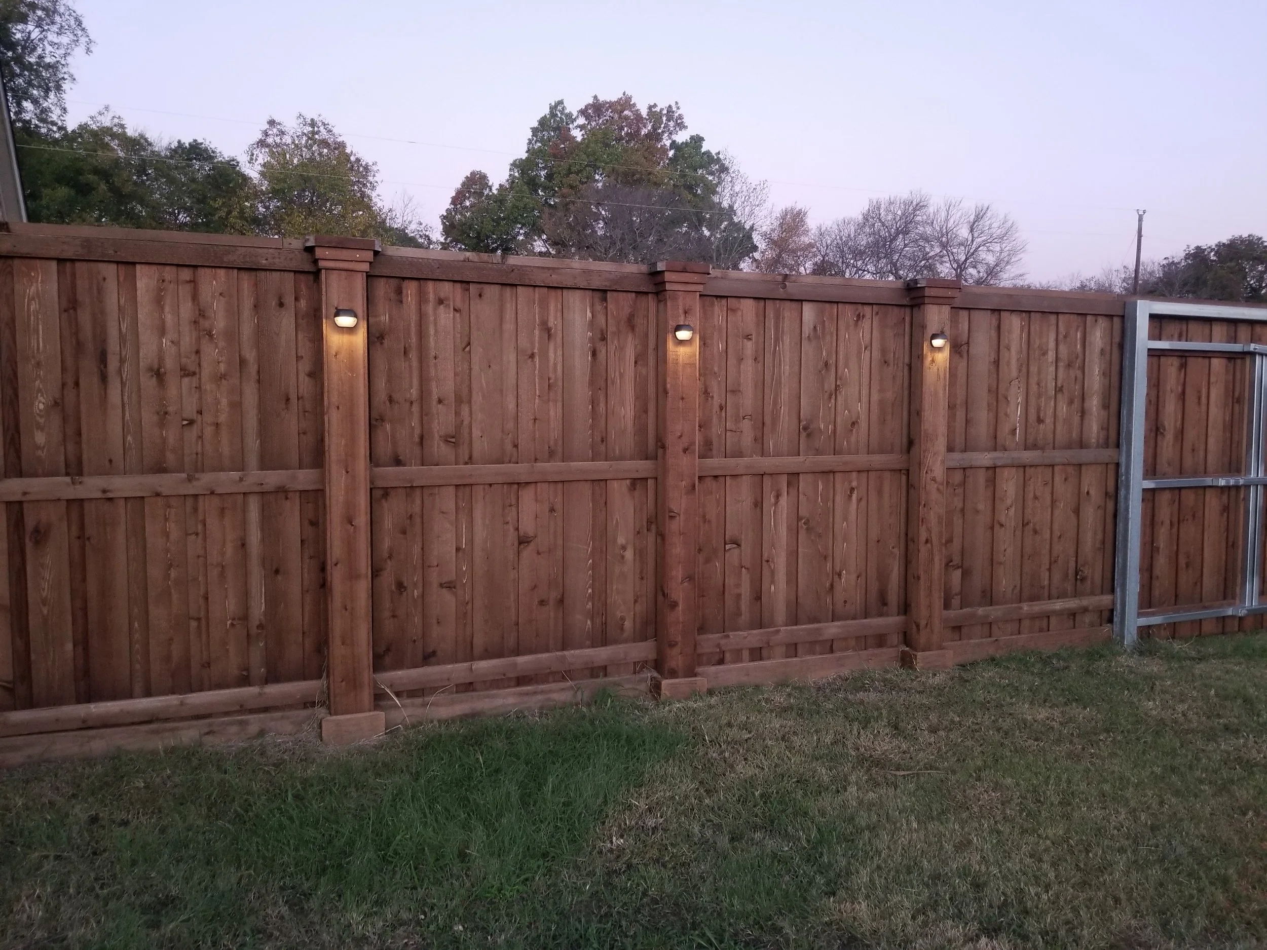 Wooden privacy fence with outdoor lights on posts, grass in the foreground, and trees in the background during dusk.