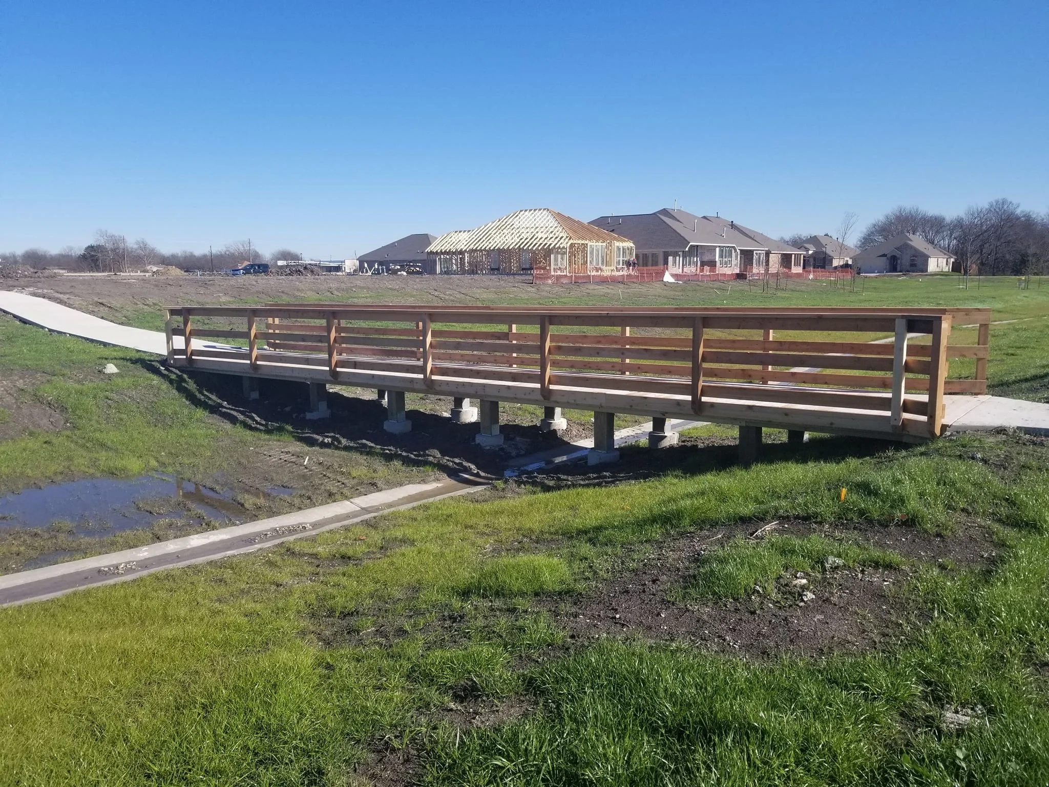 Wooden bridge over a small water drainage, construction site with houses in background on a sunny day.