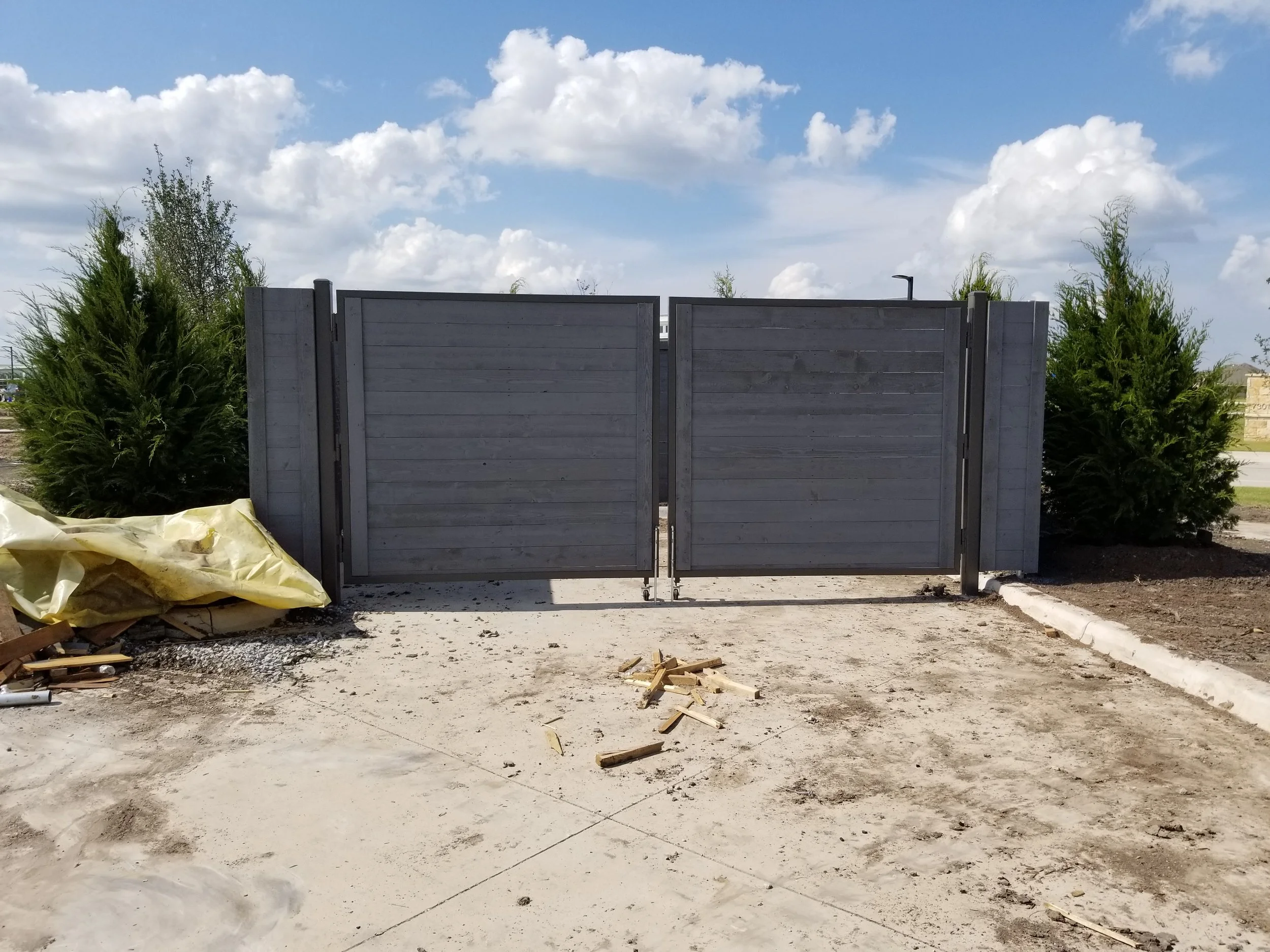 A wooden gate with metal hinges, flanked by green bushes, on a partially paved area with construction debris.