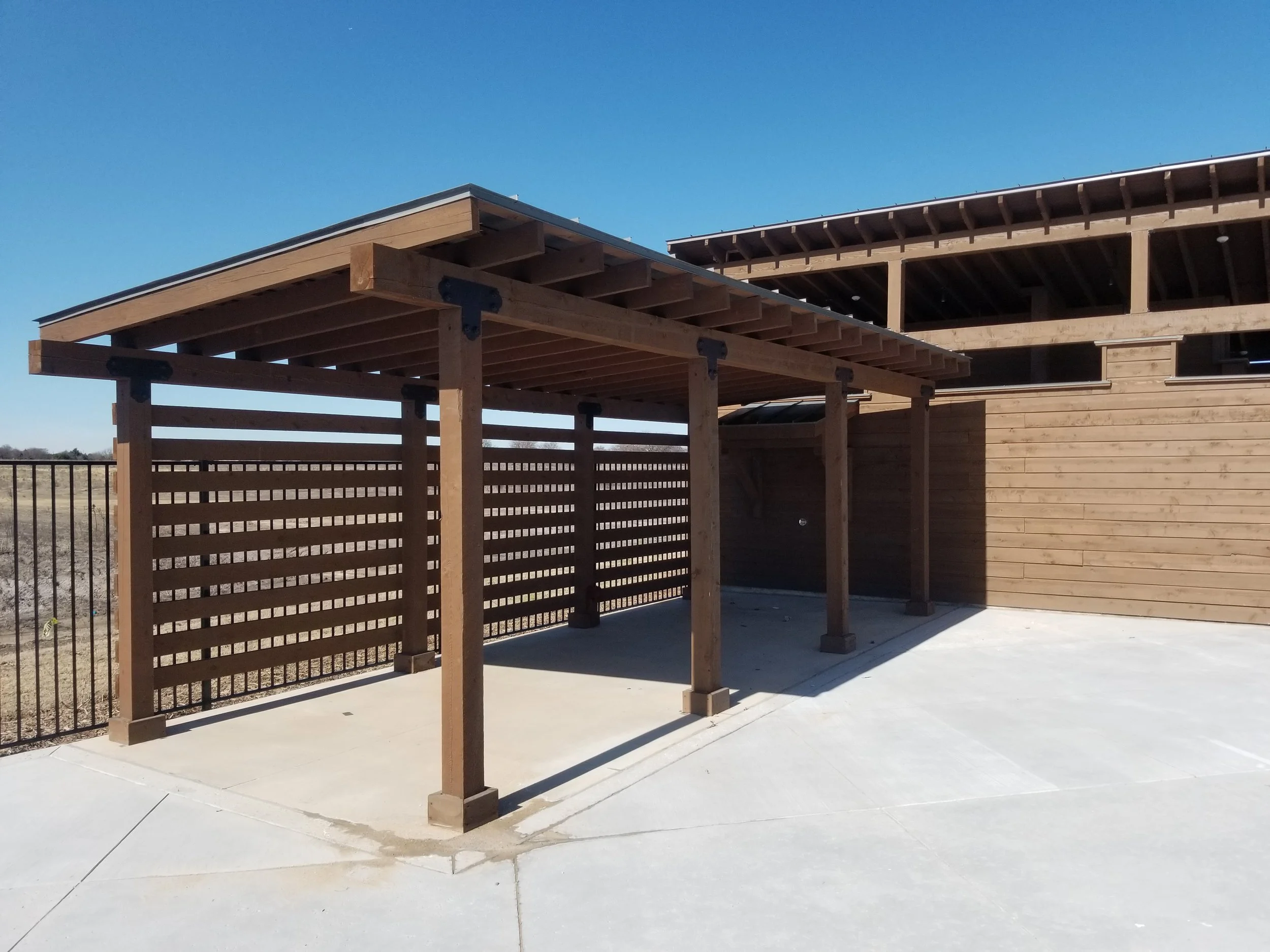 Wooden outdoor structure with a slatted fence, situated on a concrete patio under a clear blue sky, with part of a building under construction in the background.