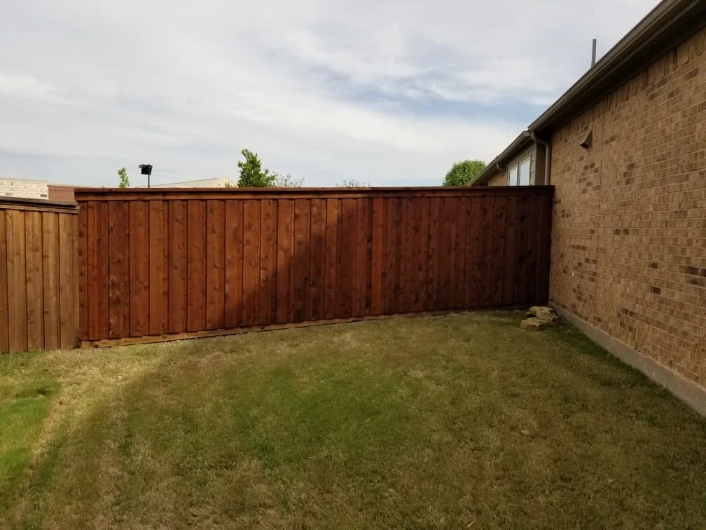 A backyard with a newly installed wooden fence, a brick house wall on the right, and a grassy yard.