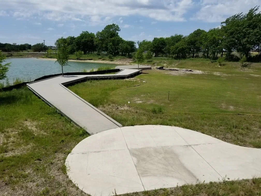 Concrete walking path and ramp leading to a lakeside area with trees and grassy fields, under a partly cloudy sky.