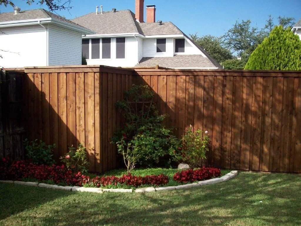 A backyard with a wooden privacy fence, green grass, and a flower bed with red flowers and bushes.