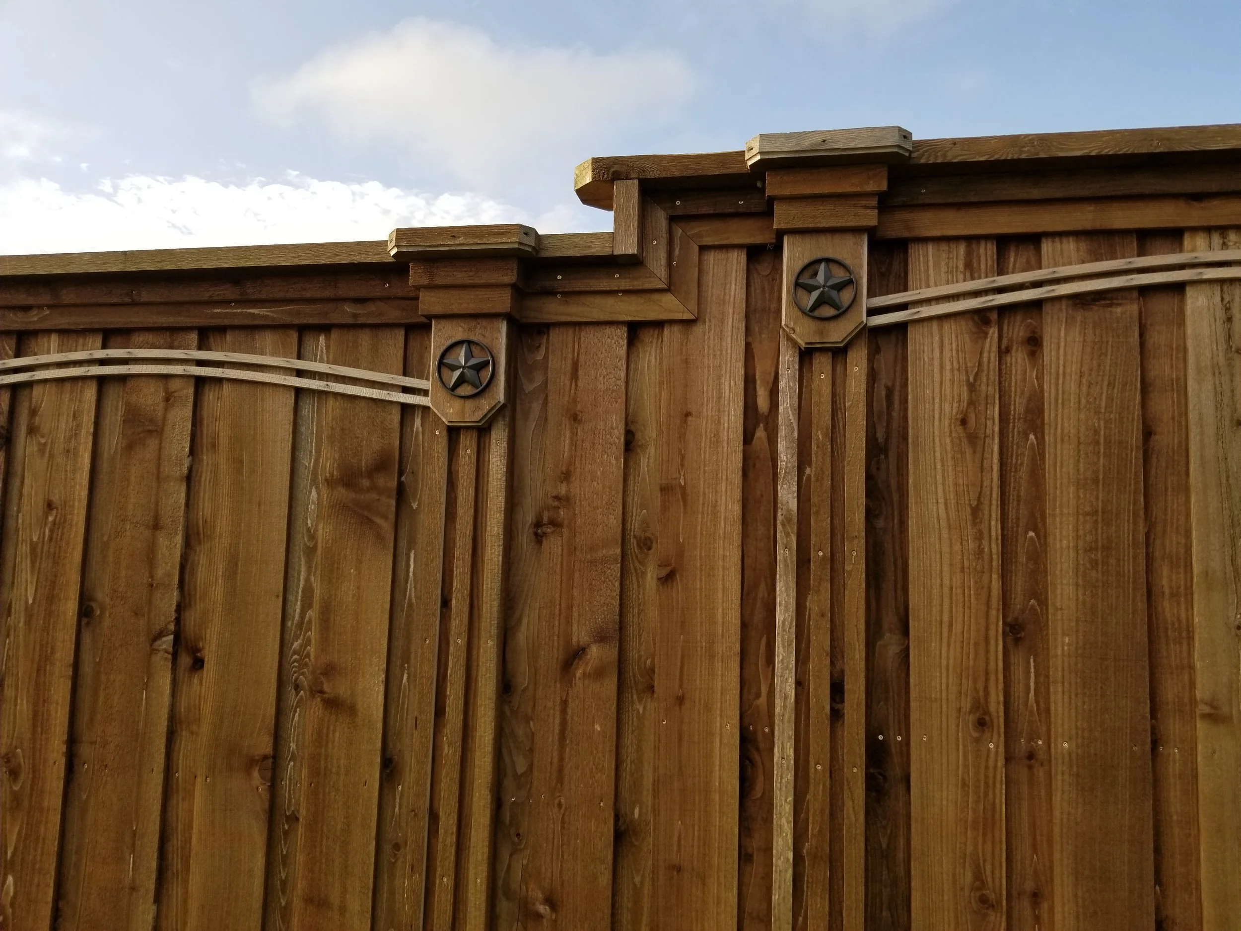 Wooden privacy fence with decorative star-shaped hardware, showing the top of the fence against a partly cloudy sky.
