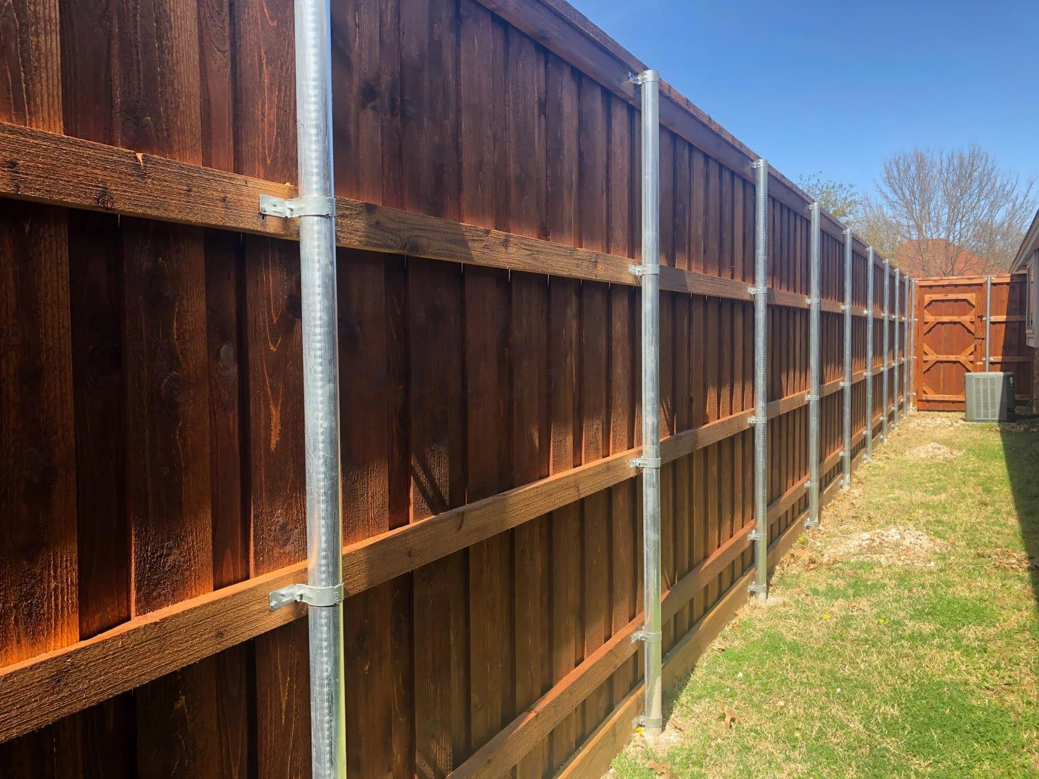 A newly installed wooden privacy fence supported by metal posts in a backyard with green grass and a clear blue sky.