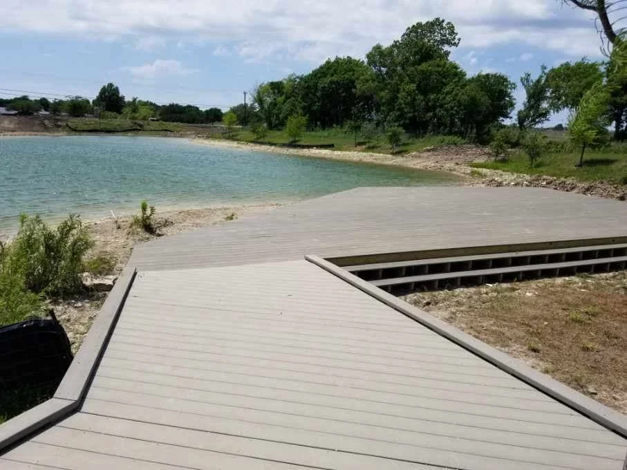 A partially completed wooden dock or platform extends over a body of water, with green trees and shrubs in the background and a cloudy sky above.