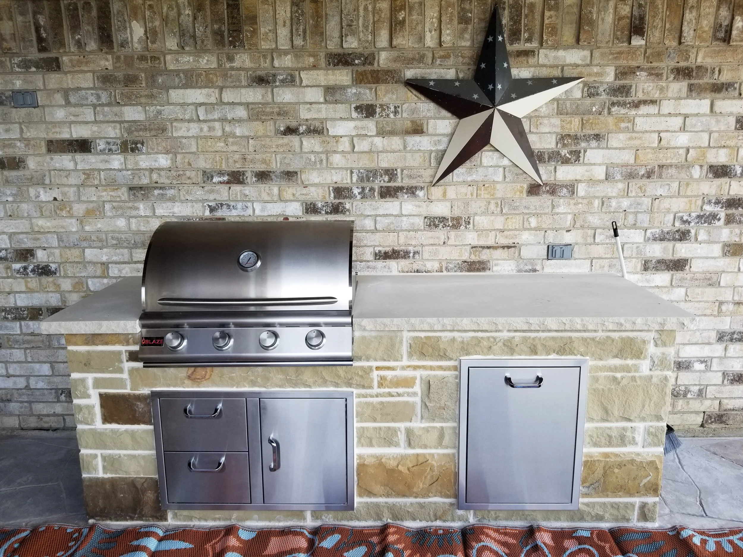 Stainless steel outdoor kitchen island with a grill, storage drawers, and cabinets against a brick wall. Two decorative stars are on the wall, and an umbrella is hidden behind the island.