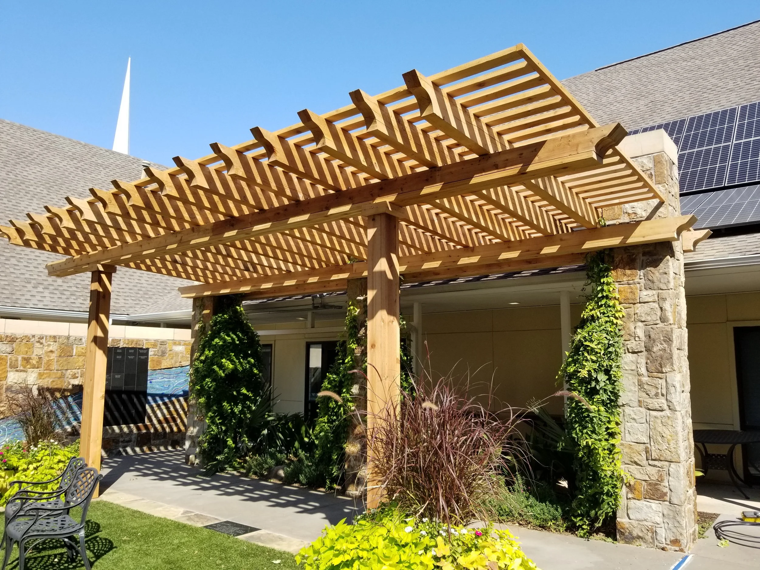 New wooden pergola with slatted roof over patio area, surrounded by plants and outdoor furniture, with solar panels on neighboring roof in the background.