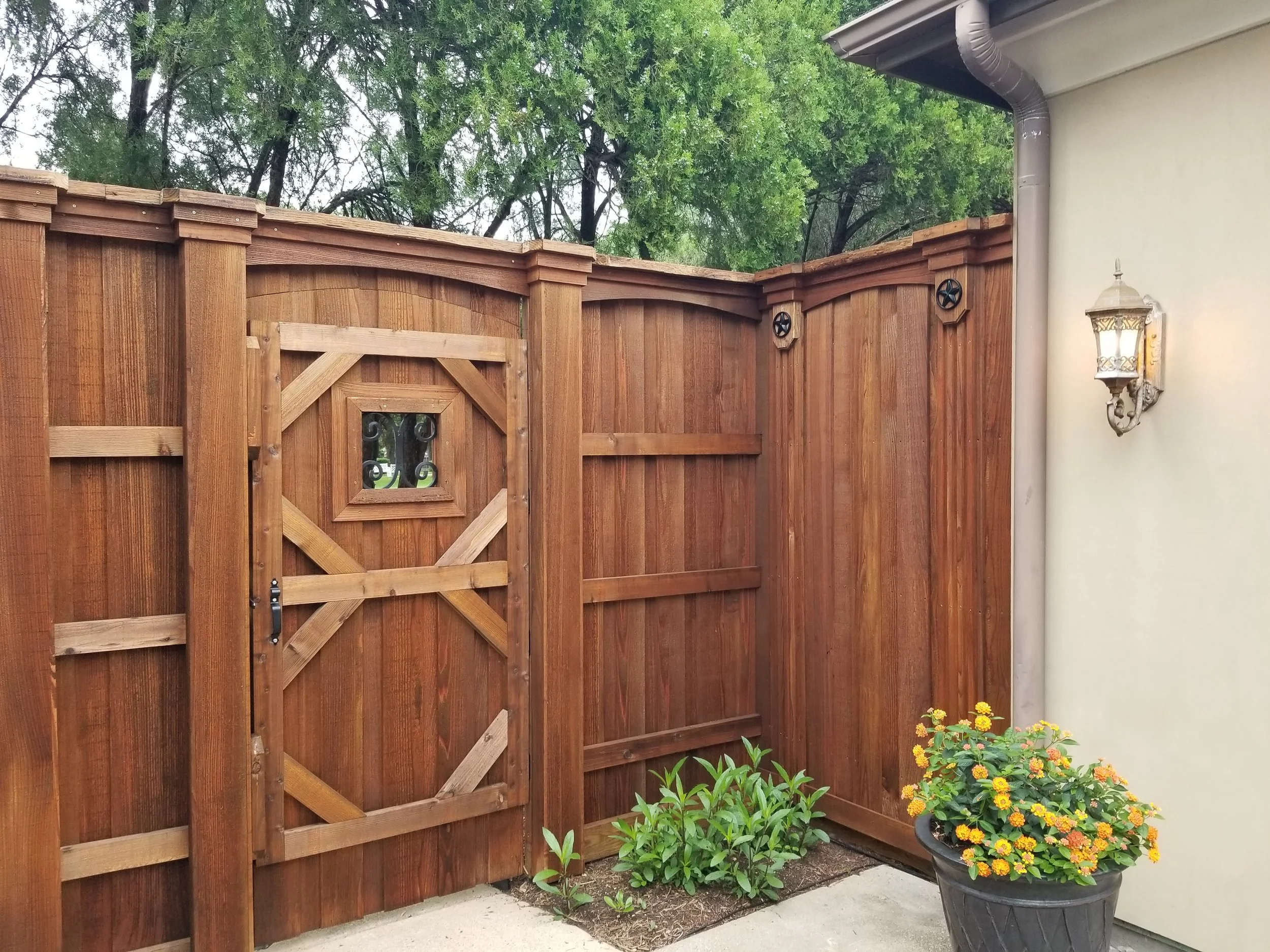 Wooden privacy fence with a small door, adjacent to a house wall with a decorative lantern and potted yellow flowering plant.