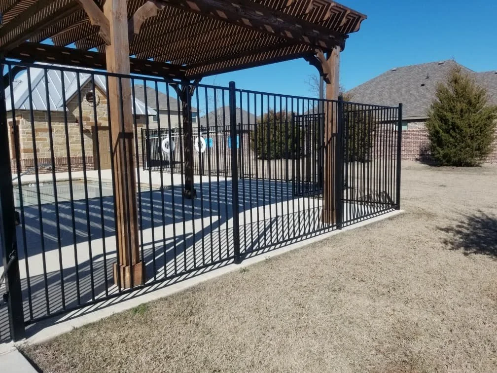A fenced outdoor patio with a wooden pergola and a concrete floor, surrounded by a lawn and neighboring houses in the background.