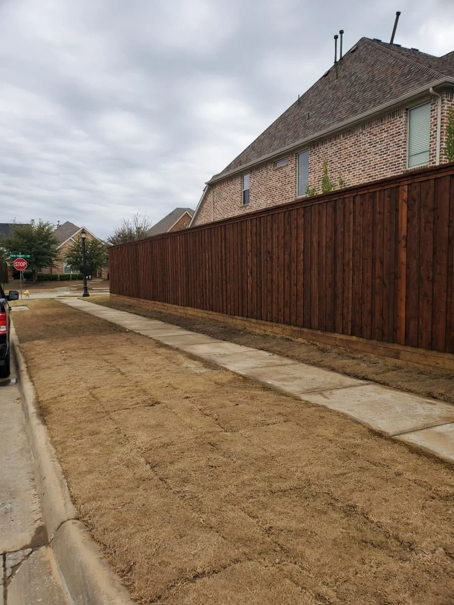 A sidewalk runs alongside a house with a tall wooden fence. The house is made of brick, and the sky is overcast.