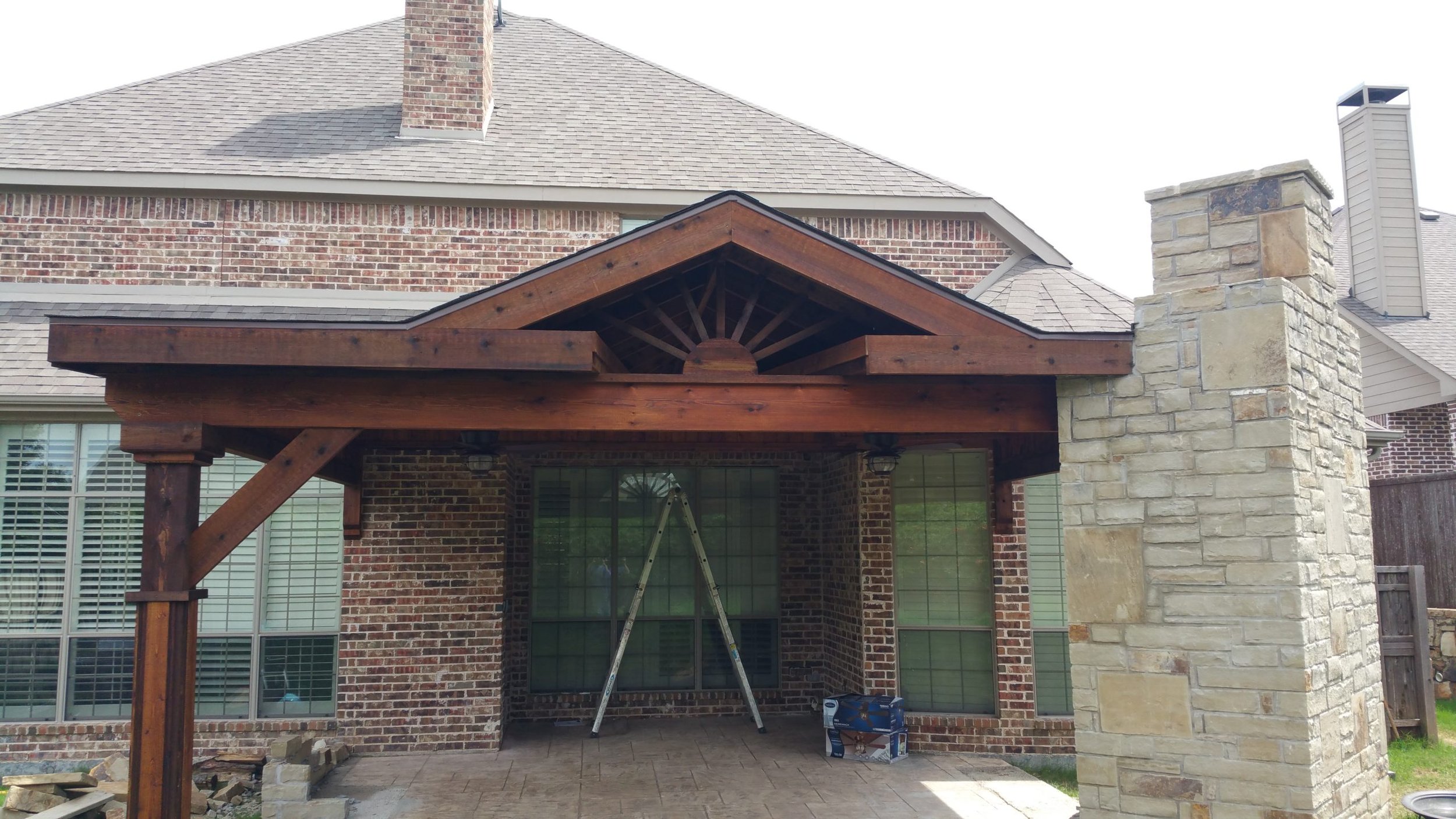 Back patio of a brick house with a wooden porch roof, a stone chimney, large windows, and a ladder set up in front of the windows, possibly for work or repair.