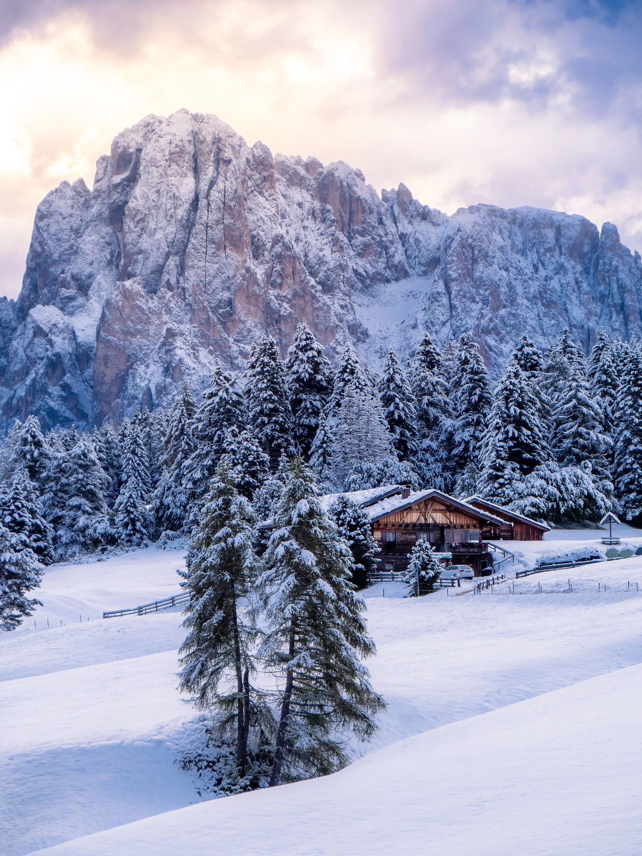Winter auf der Seiser Alm: Unberühte Schneedecke, verschneite Tannen und dazwischen eine Almhütte. Im Hintergrund erhebt sich majestätisch der Langkofel.