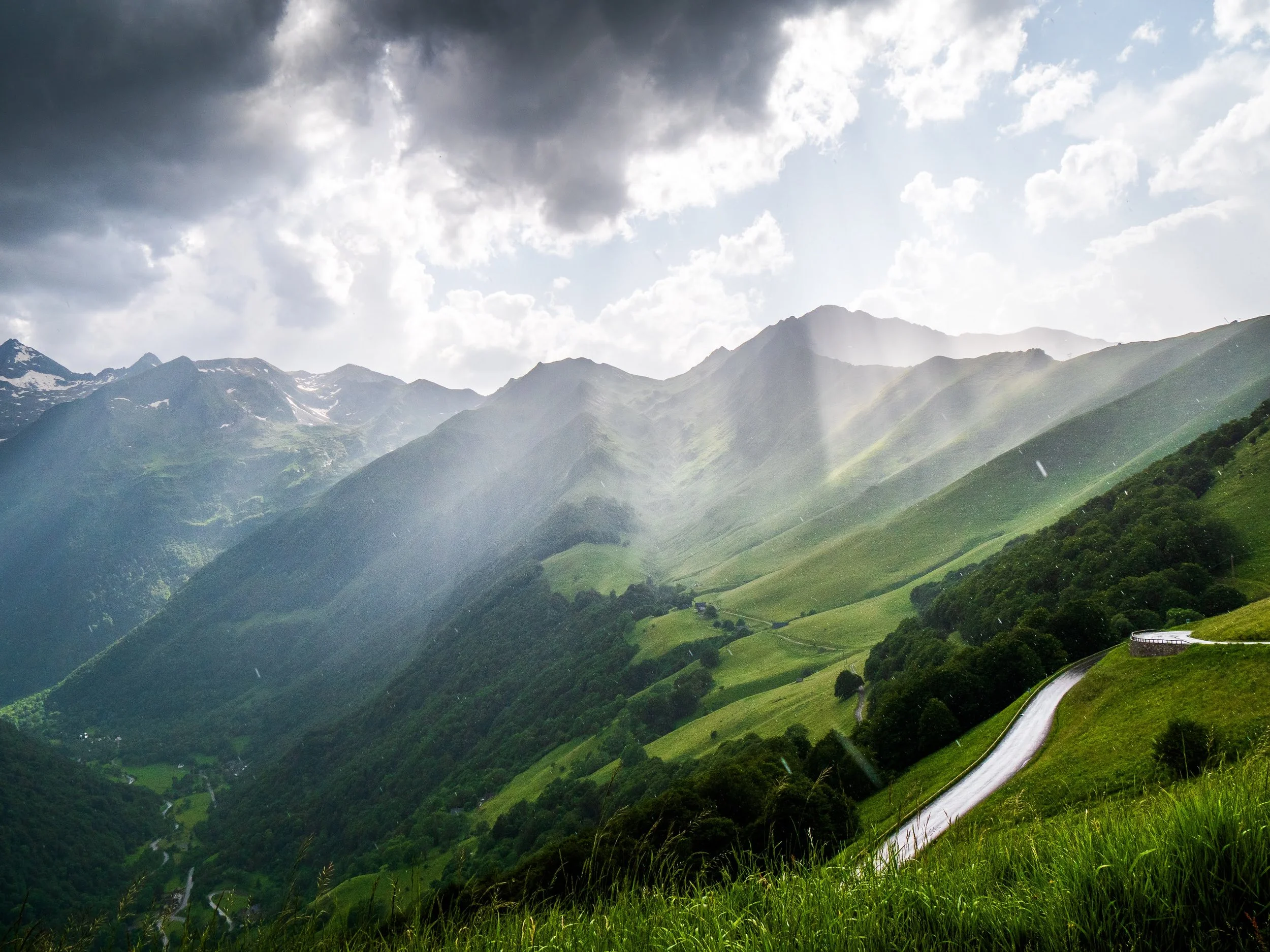 Wetterstimmung in den Pyrenäen auf einer Passstraße: Aussicht über mehrere Berge. Sonnenstrahlen leuchten durch dunkle Gewitterwolken, fallende Regentropfen sind zu erkennen. Eine regennasse Straße glitzert im Vordergrund.