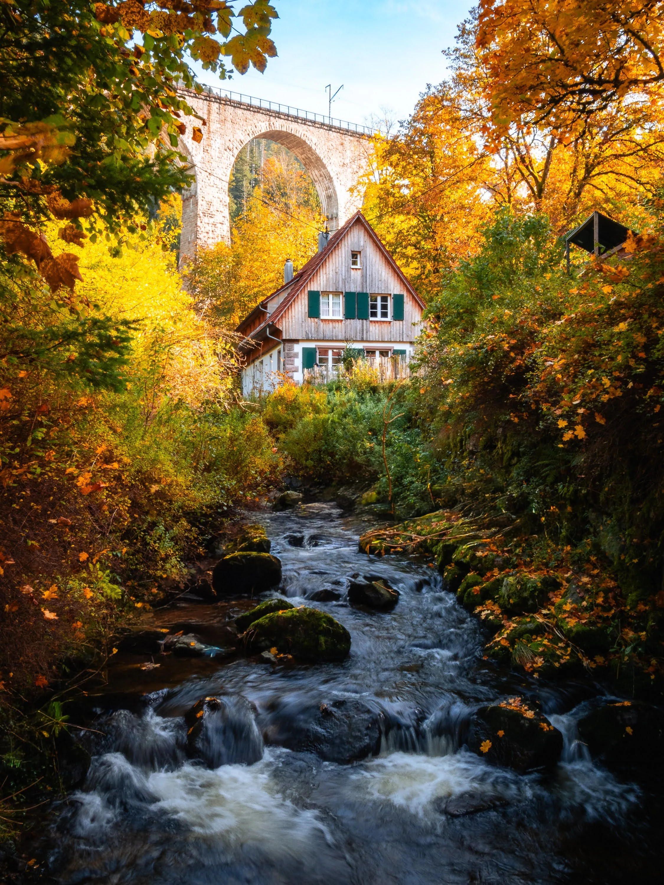 Die Ravenna-Lodge bei Breitnau: Ein Haus steht an einem malerischen Bach, im Hintergrund ein Viadukt. Herbstliche Szene mit buntem Laub und blauem Himmel.