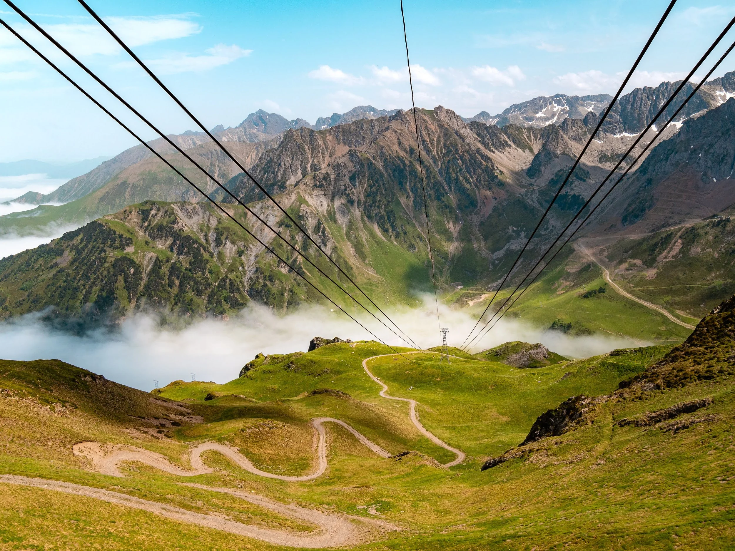 Panoramablick aus der Gondel am Pic du Midi de Bigorre: Eine gewundene Straße, die Mittelstation, dahinter Nebelschwaden und Berge