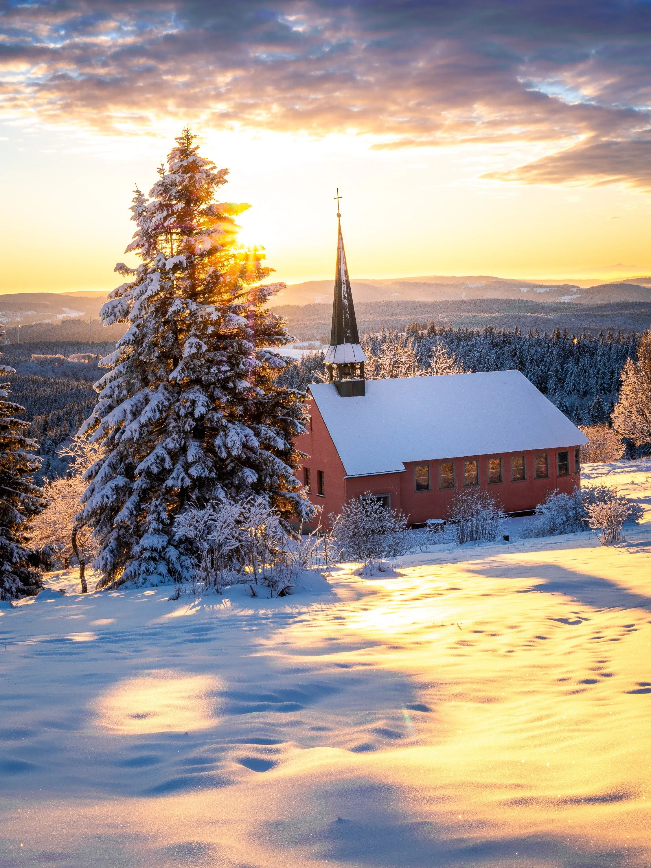 Goldener Wintermorgen-Moment: Zwischen einer verschneiten Tanne und der kleinen Kandelkapelle St. Pius geht die Sonne auf.