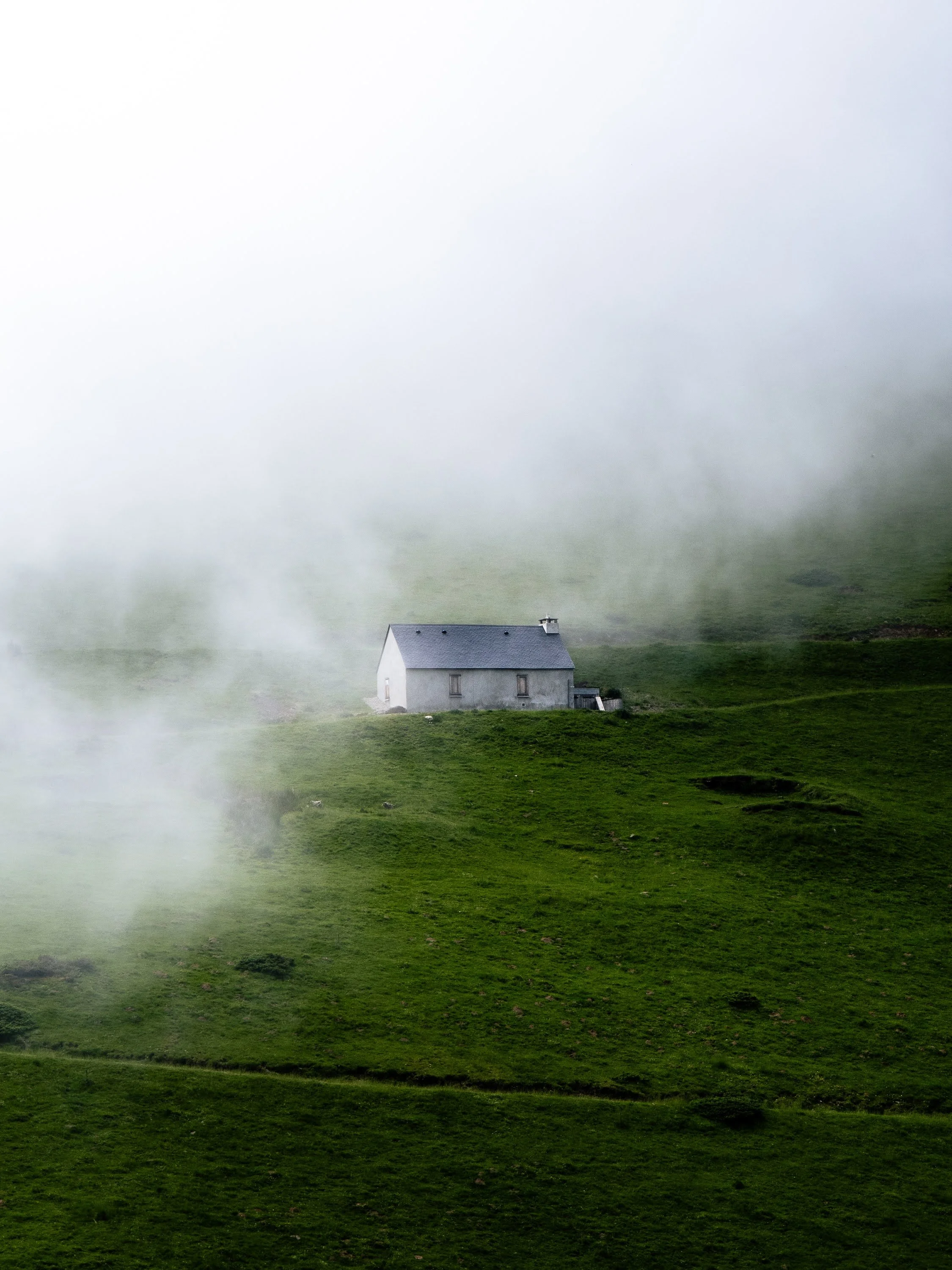 Ein einsames kleines Haus an einem grünen Hang. Stimmungsvoller Nebel bedeckt die obere Bilddiagonole, sodass nur das Haus zu sehen ist.