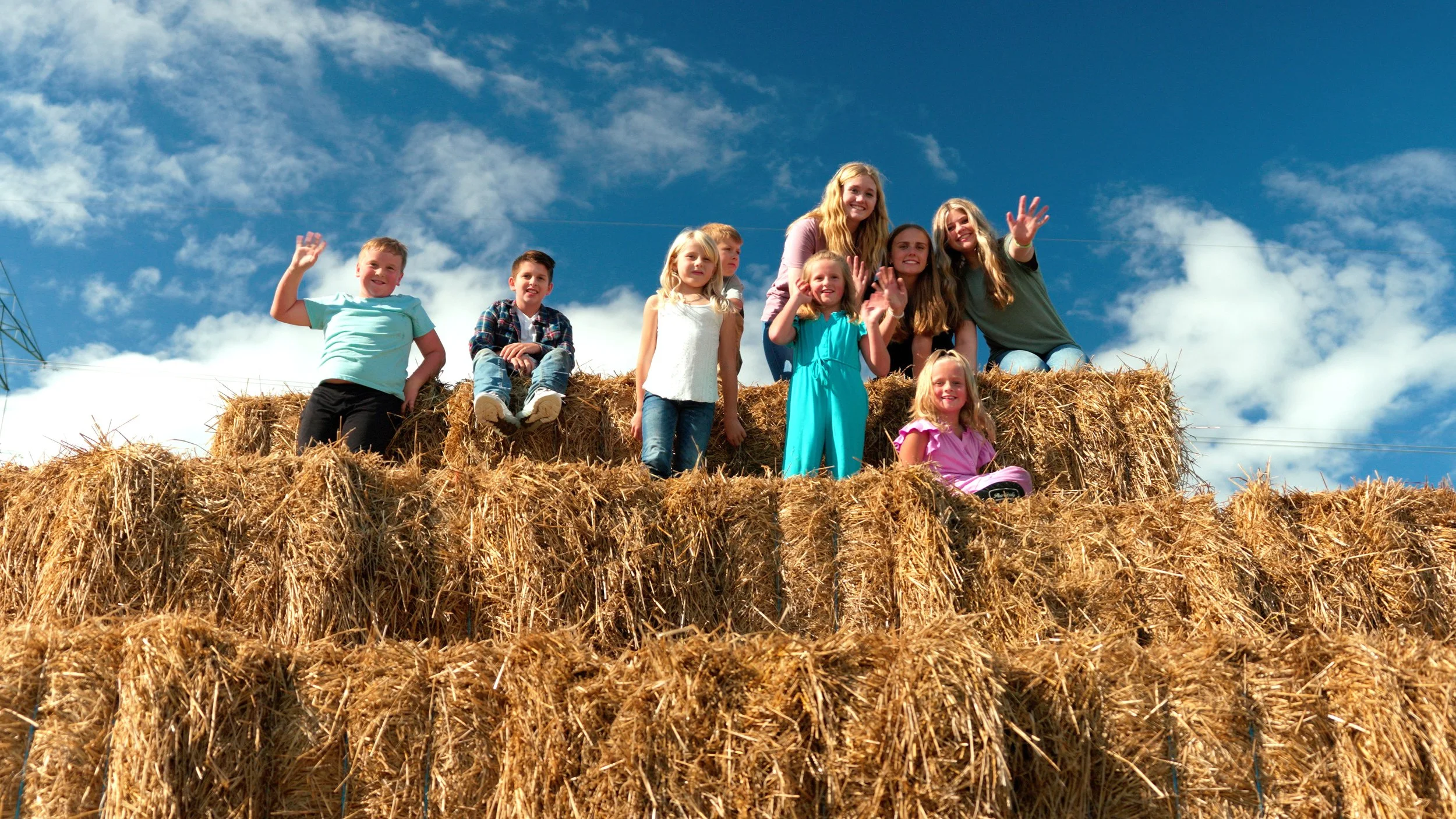 Children and adults sitting on top of hay bales outdoors, smiling and waving, against a bright blue sky with some clouds.