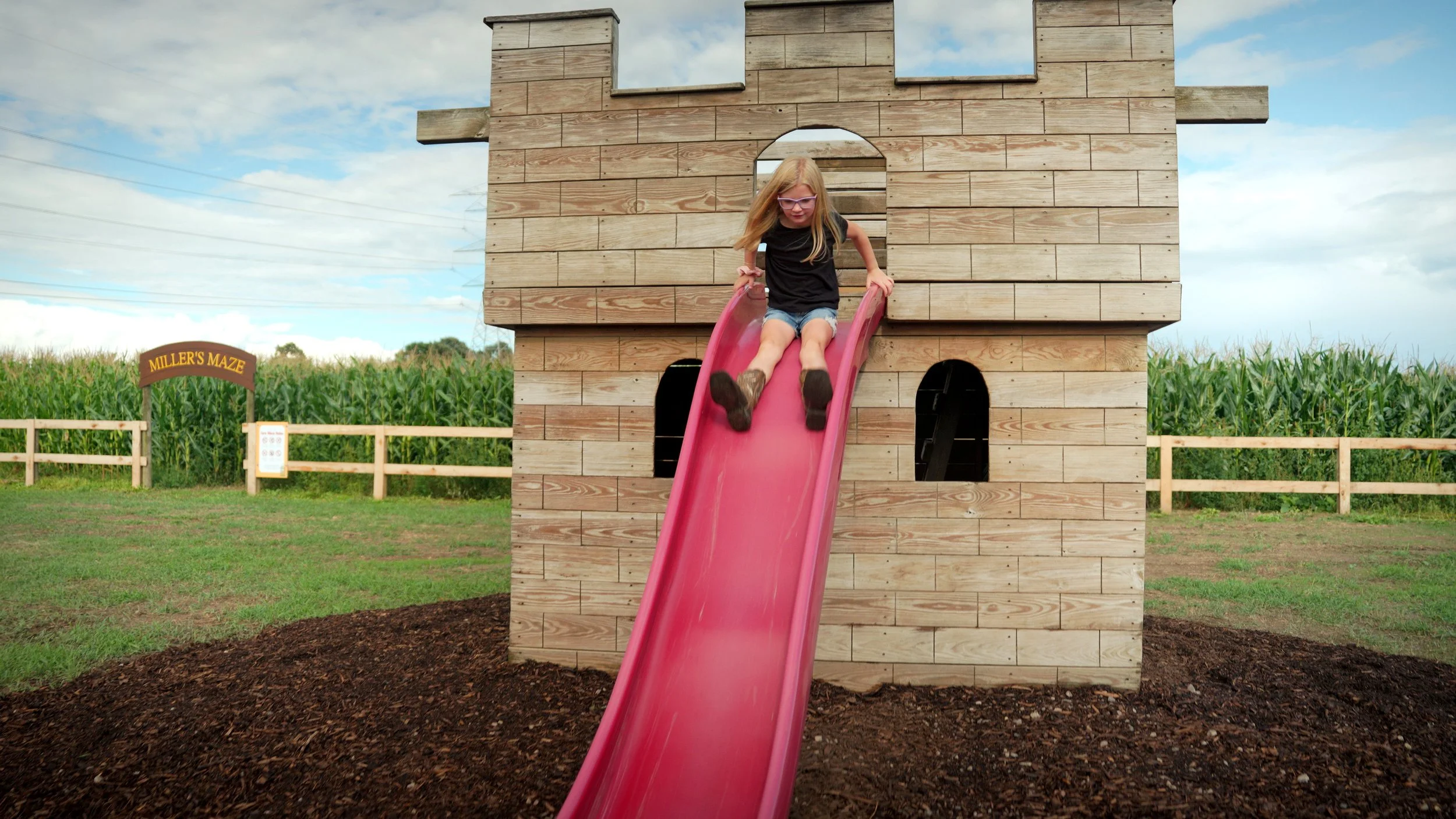 A young girl with long blonde hair, wearing glasses, a black shirt, and denim shorts, is sliding down a pink slide attached to a wooden playhouse shaped like a castle in an outdoor play area with grass, a wooden fence, and green cornfield in the back