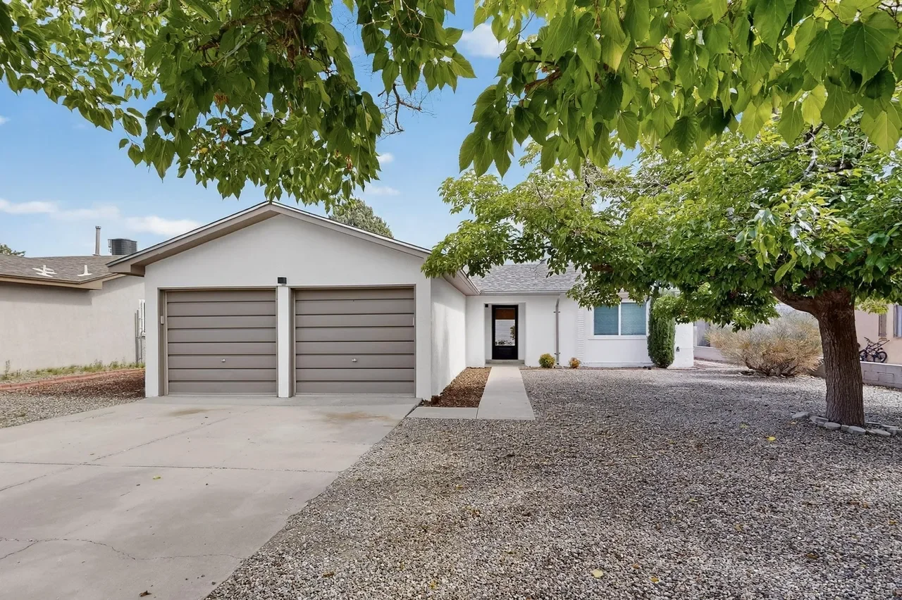 Front view of a modern house with a two-car garage, concrete driveway, and gravel yard, partially shaded by large green trees, in a suburban neighborhood.