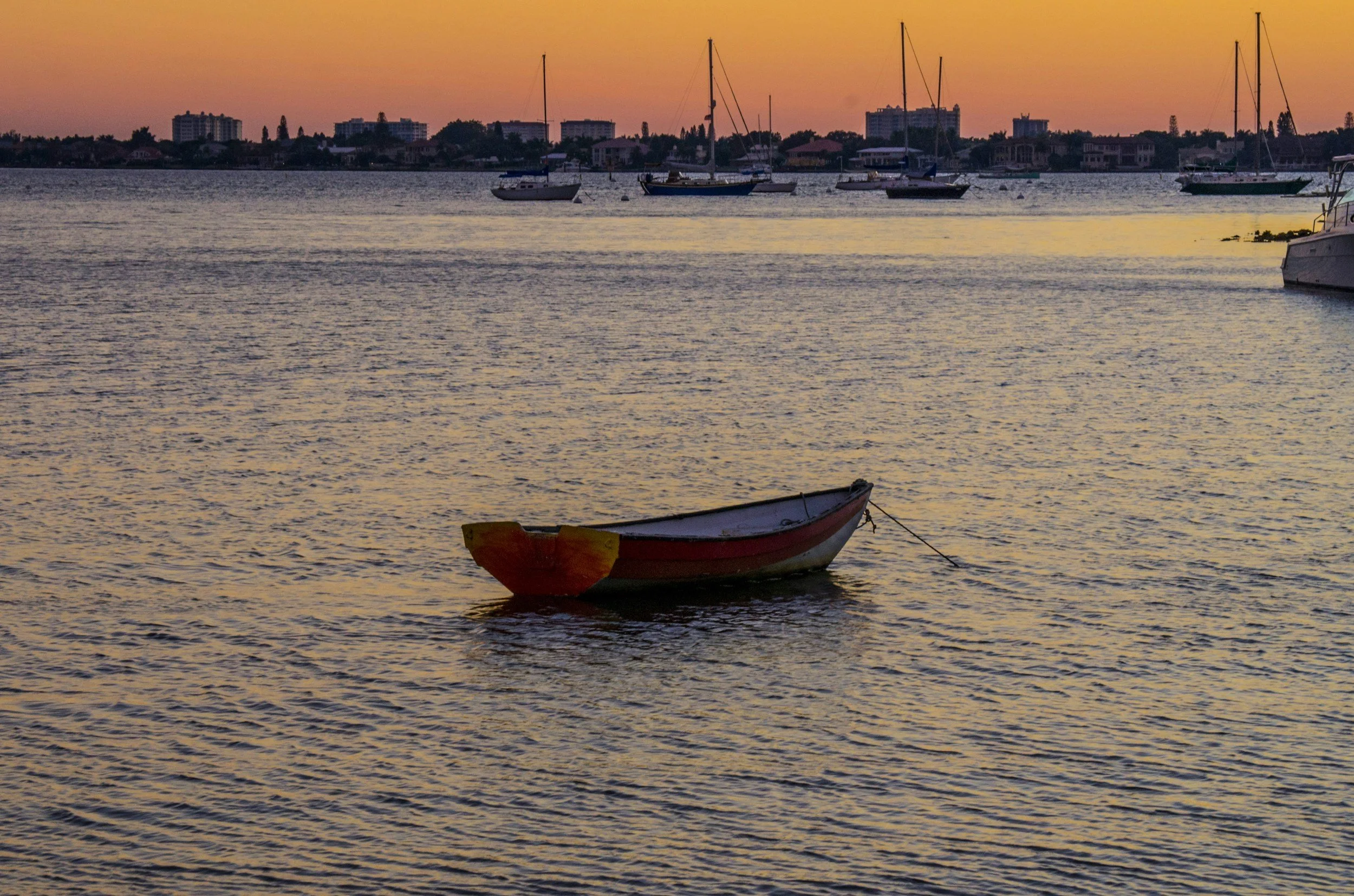 A small boat floating on calm water during sunset, with sailboats and city buildings in the background.
