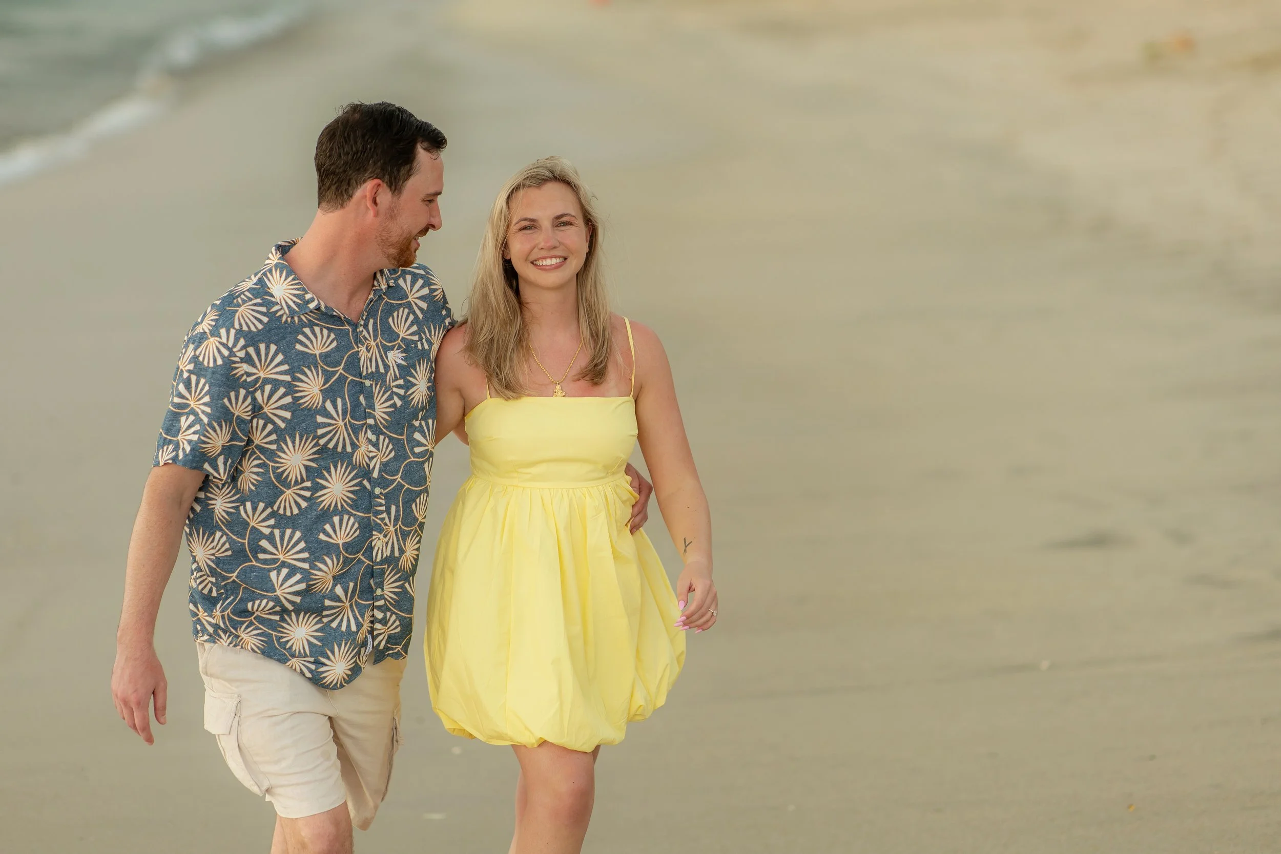 A smiling couple walking along the beach, with the man wearing a blue Hawaiian shirt and white shorts, and the woman in a yellow dress, holding hands and enjoying the seaside scenery.