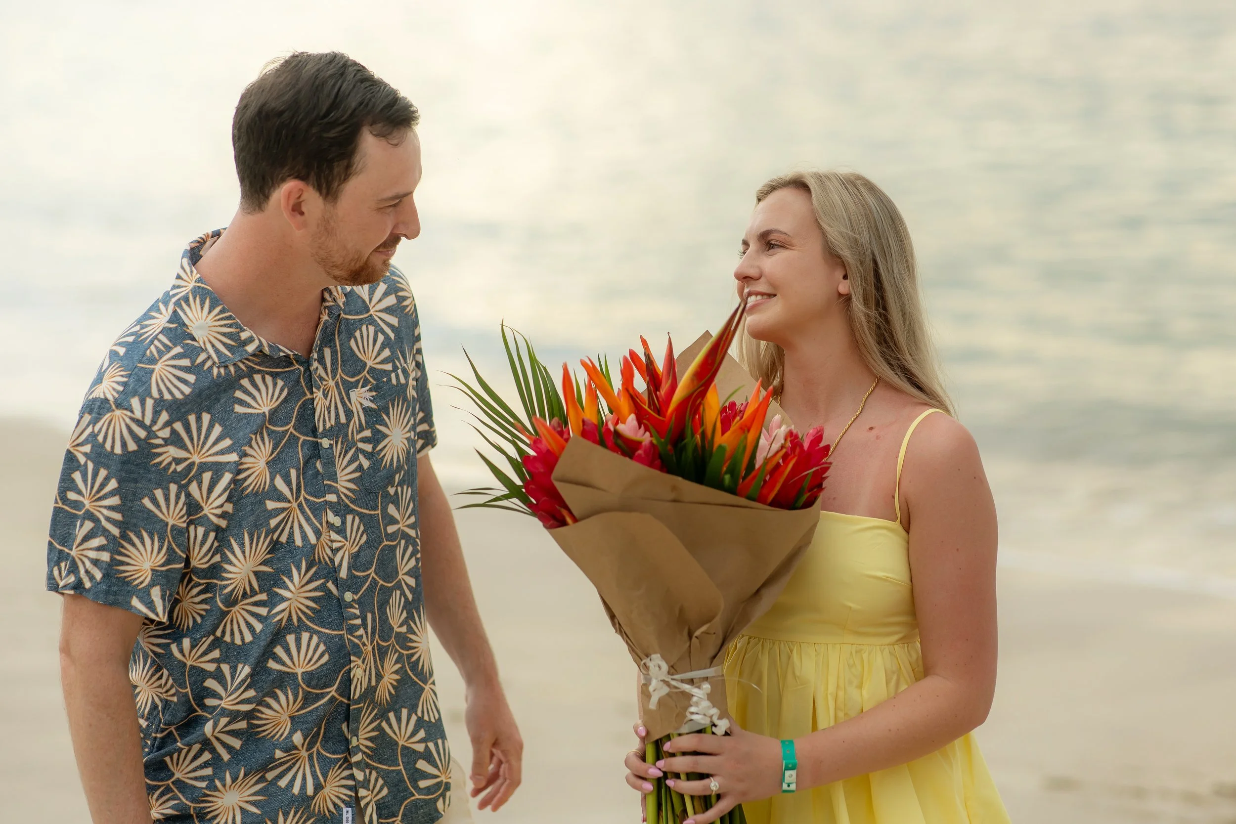 A man offering a bouquet of tropical flowers to a smiling woman on a beach.