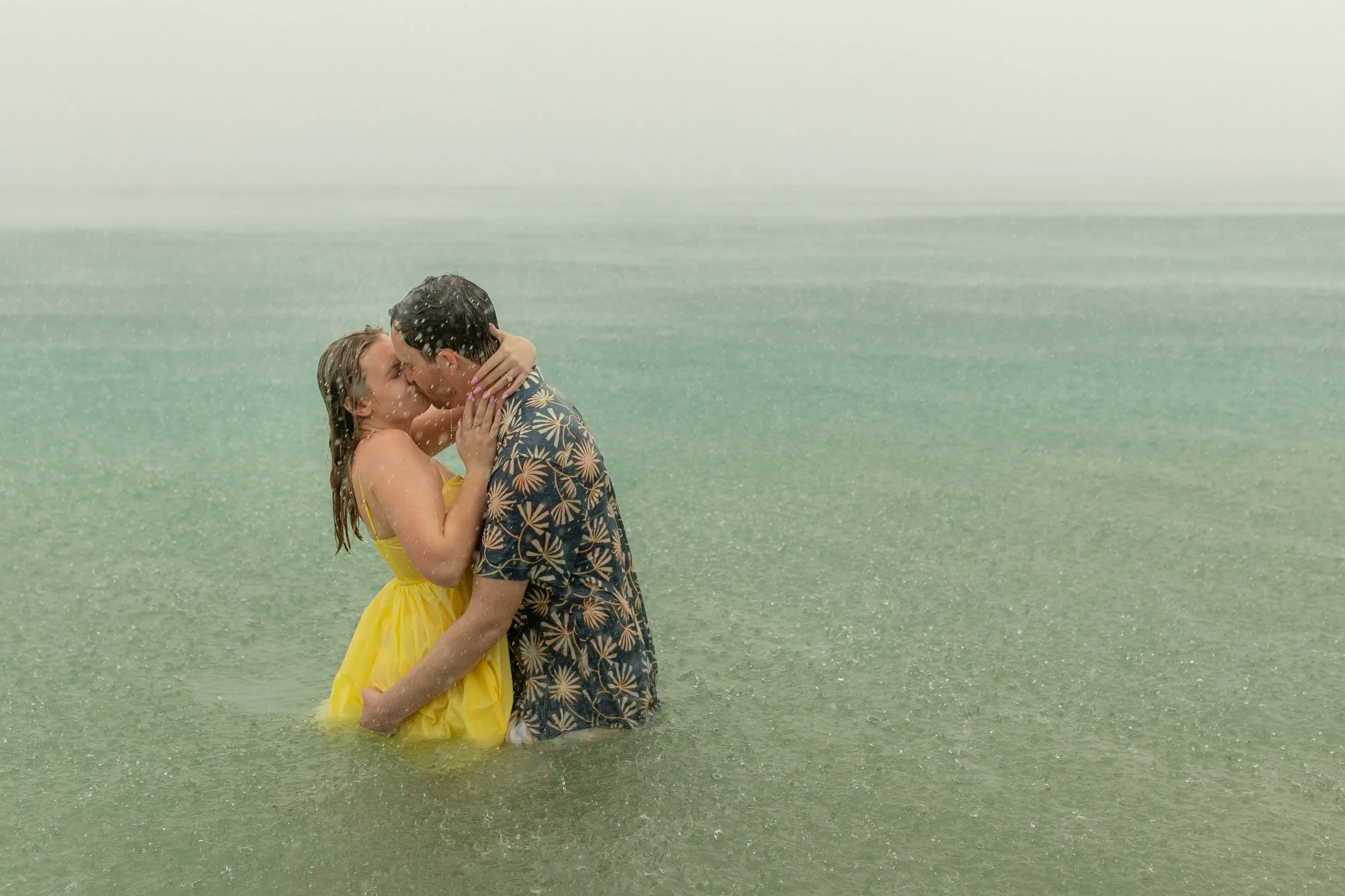 A couple sharing a kiss in the ocean during a rainstorm, with water reaching their waists.