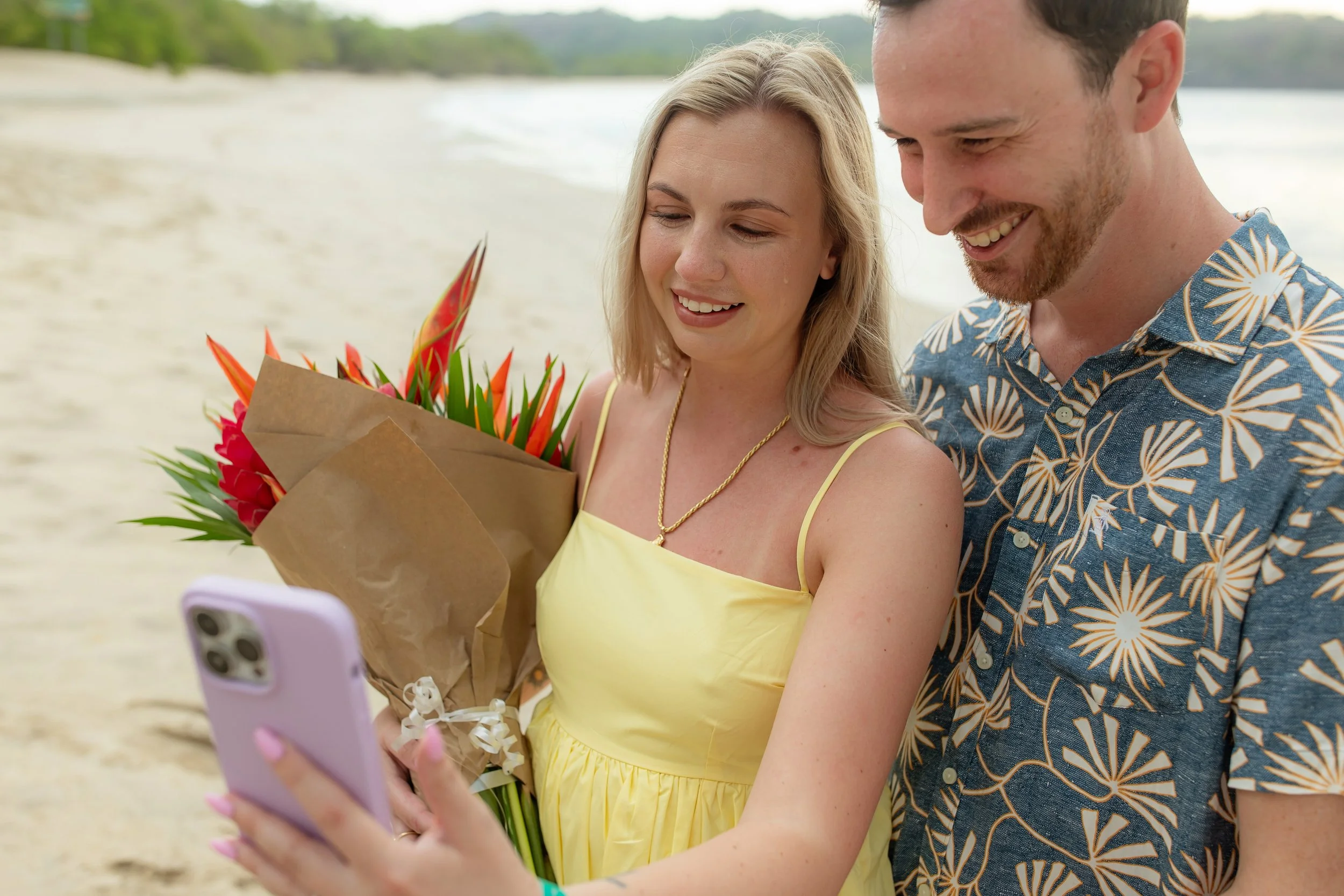 A woman in a yellow dress holding a bouquet of tropical flowers and looking at a smartphone, while a man in a Hawaiian shirt stands beside her on the beach, both smiling.