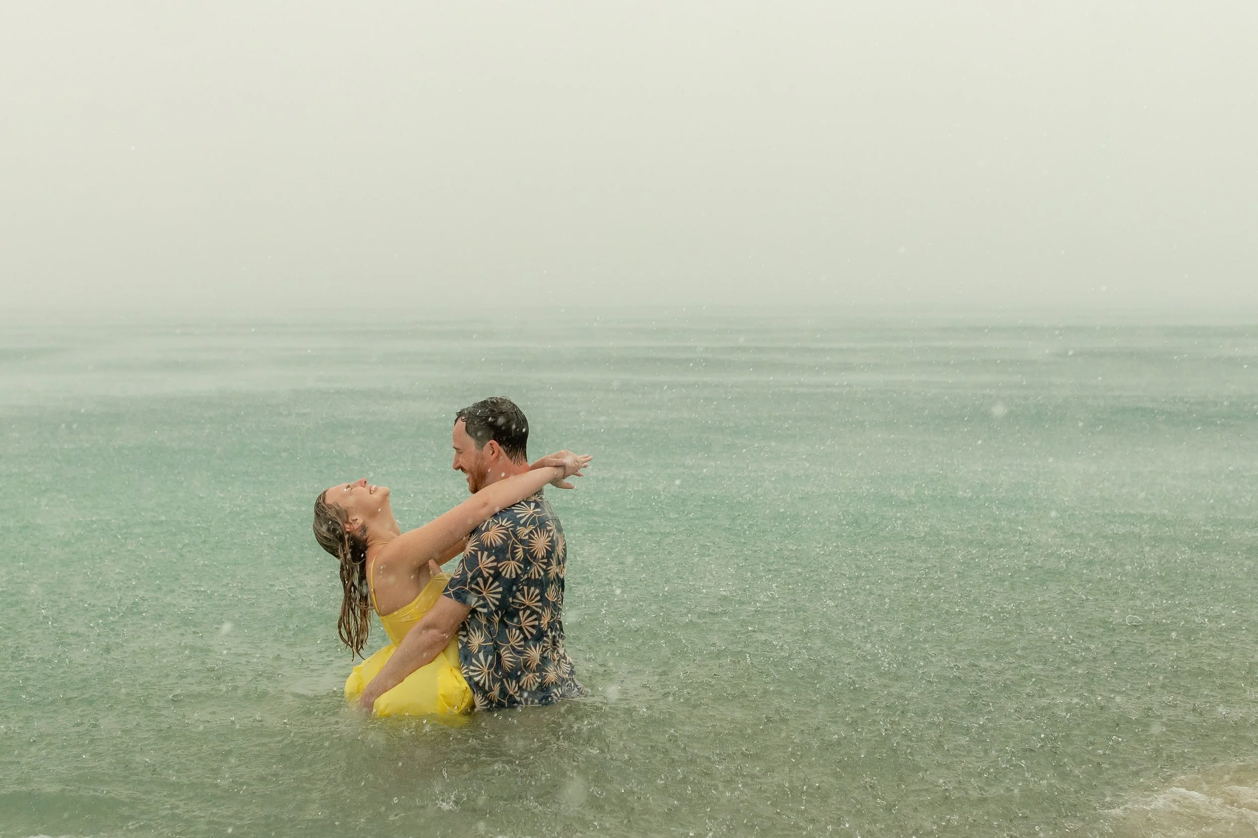 A man and a woman are in the ocean, smiling and embracing each other during a rainstorm.