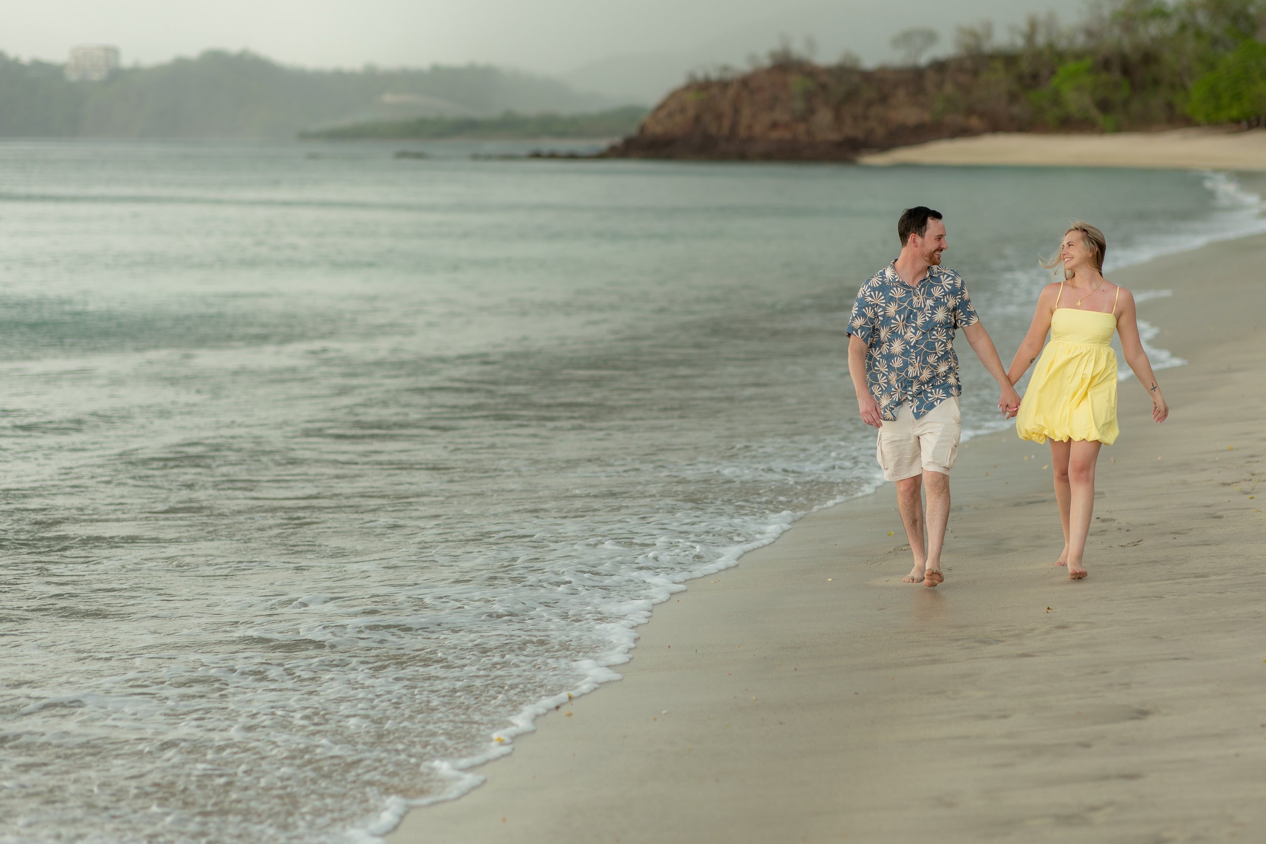 A couple holding hands and walking along the beach at sunset, with shoreline and water in the background.