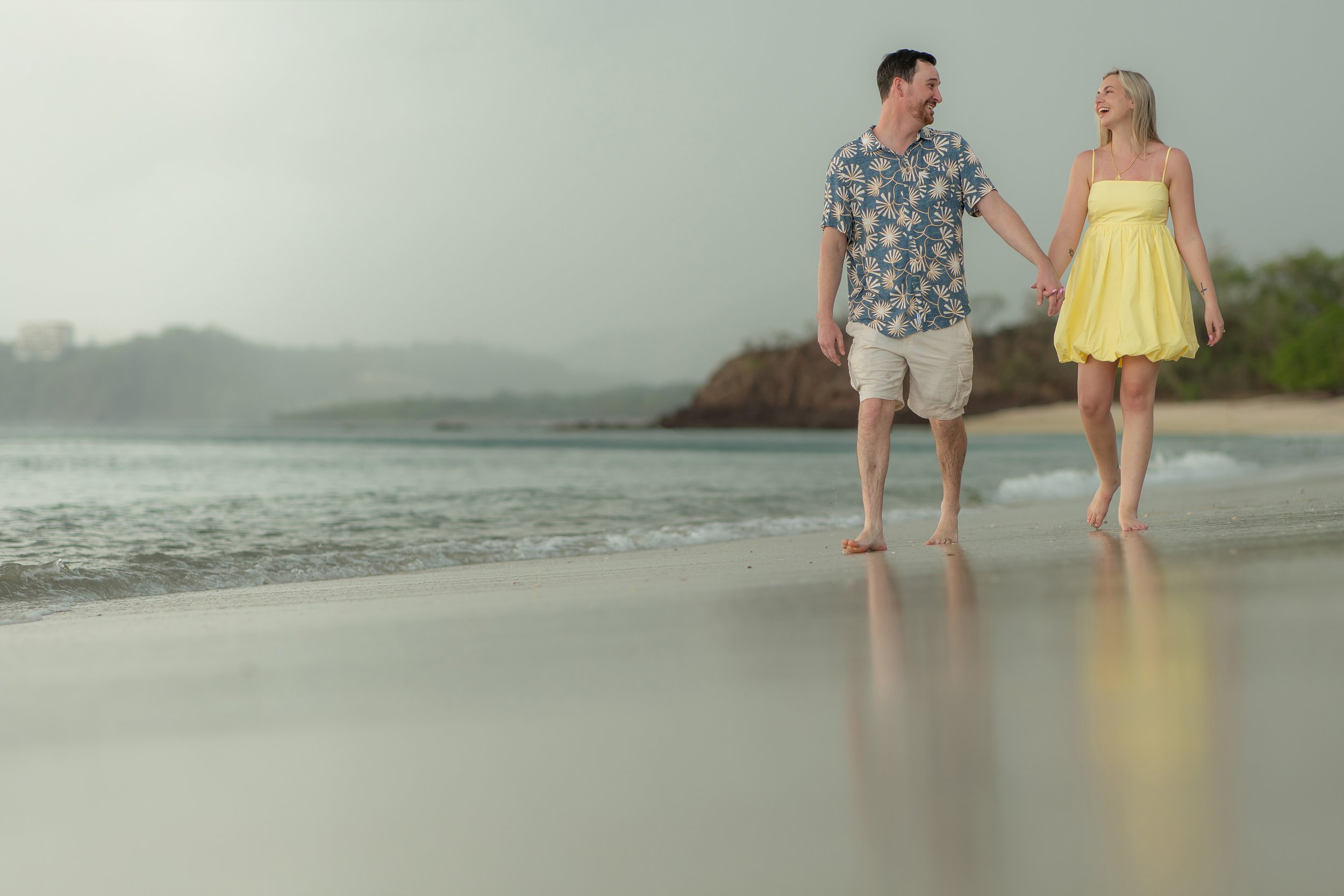 A couple is walking hand in hand along the beach near the ocean, smiling and enjoying each other's company.