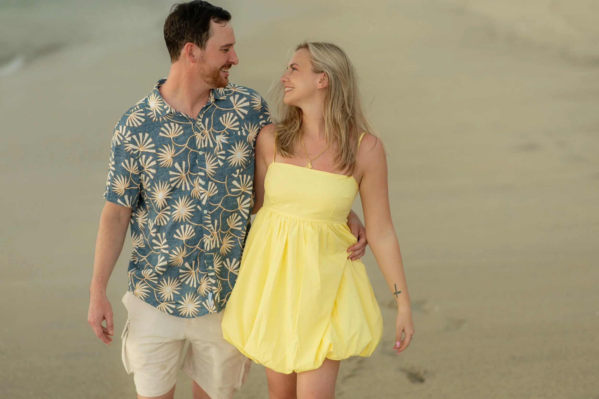 A smiling couple walking on the beach, the man is wearing a blue Hawaiian shirt with white floral patterns and beige shorts, and the woman is wearing a yellow sundress, with blond hair and a cross tattoo on her left forearm.