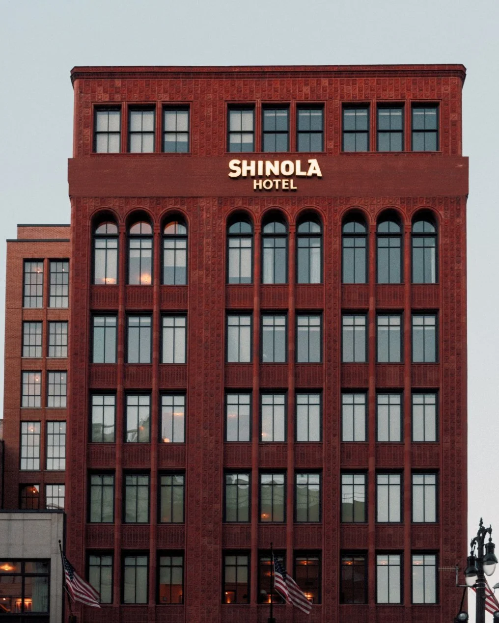 Red brick hotel building with the sign 'Shinola Hotel' at the top, multiple windows, flags in front, and decorative lamp posts nearby.