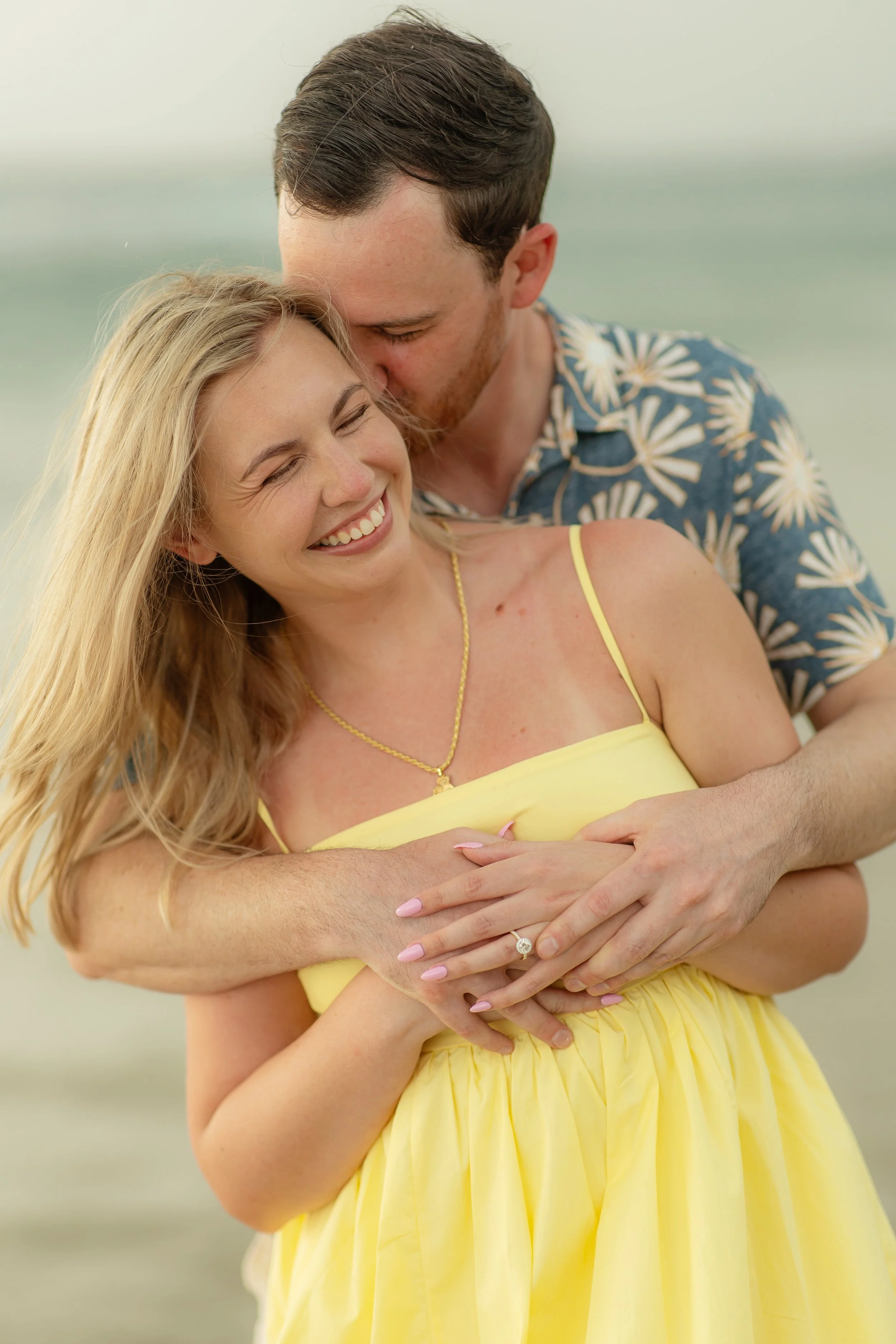 A happy couple on the beach, with the man kissing the woman on her temple as she smiles with eyes closed, wearing a yellow dress and a ring on her finger.