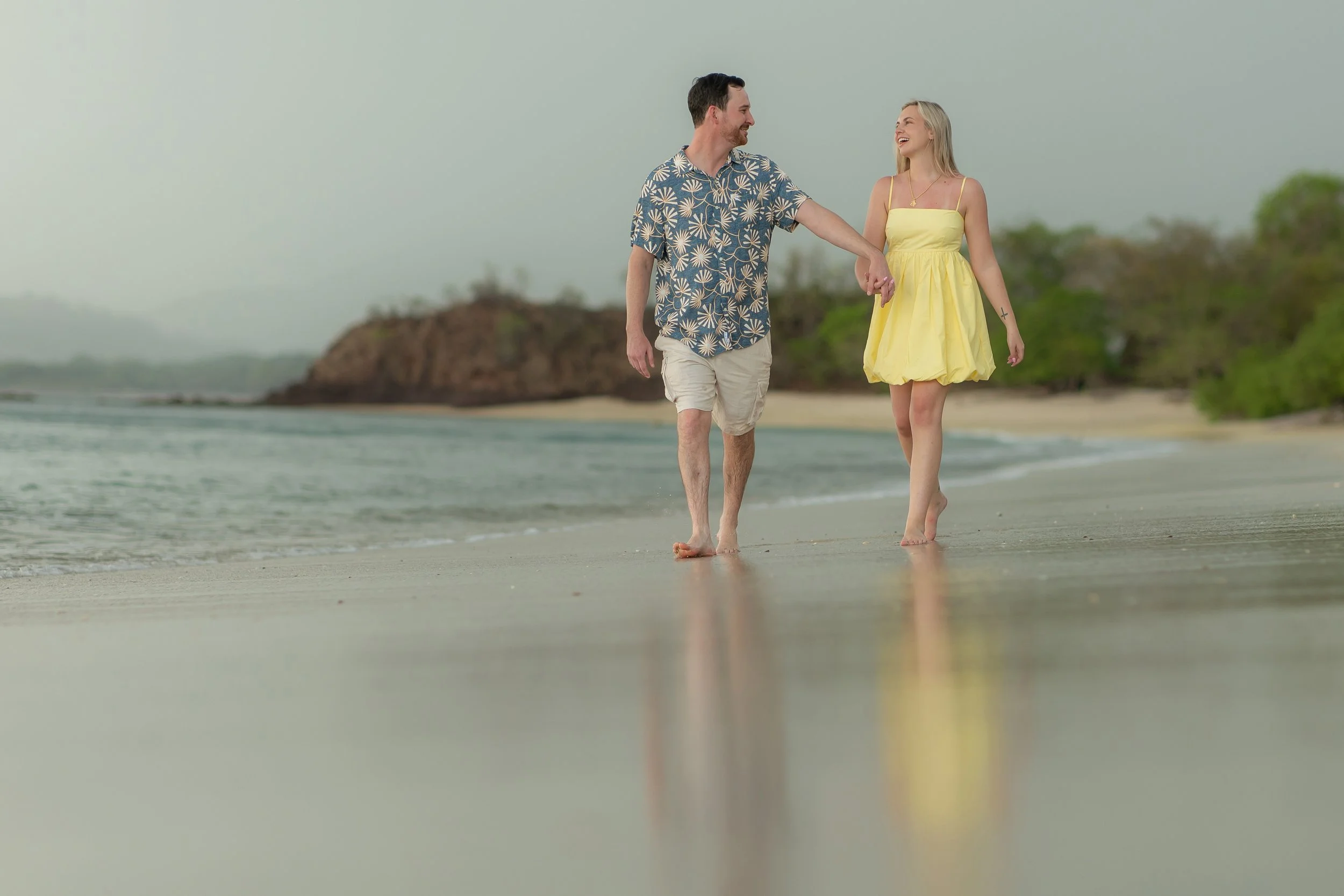 A man and woman walking hand in hand along the beach, smiling and enjoying the scenery.