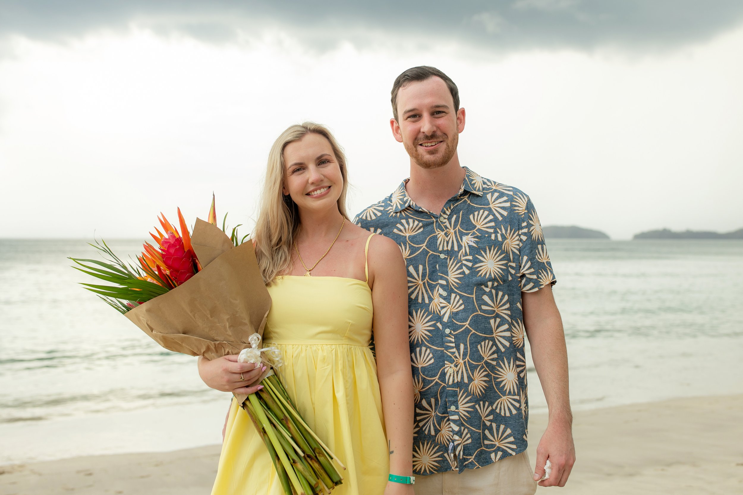 A smiling woman in a yellow dress holding a bouquet of tropical flowers standing next to a smiling man in a blue Hawaiian shirt on a beach with the ocean and distant islands in the background.