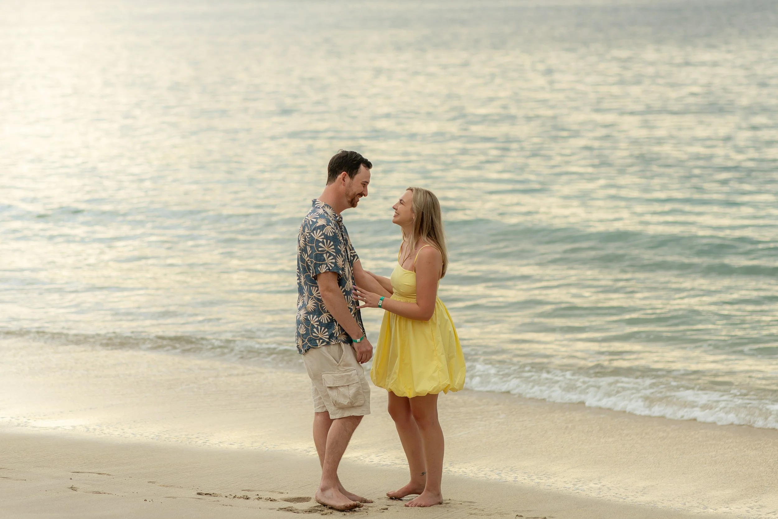 A man and woman stand close together on a beach, smiling and looking into each other's eyes with the ocean behind them during sunset.