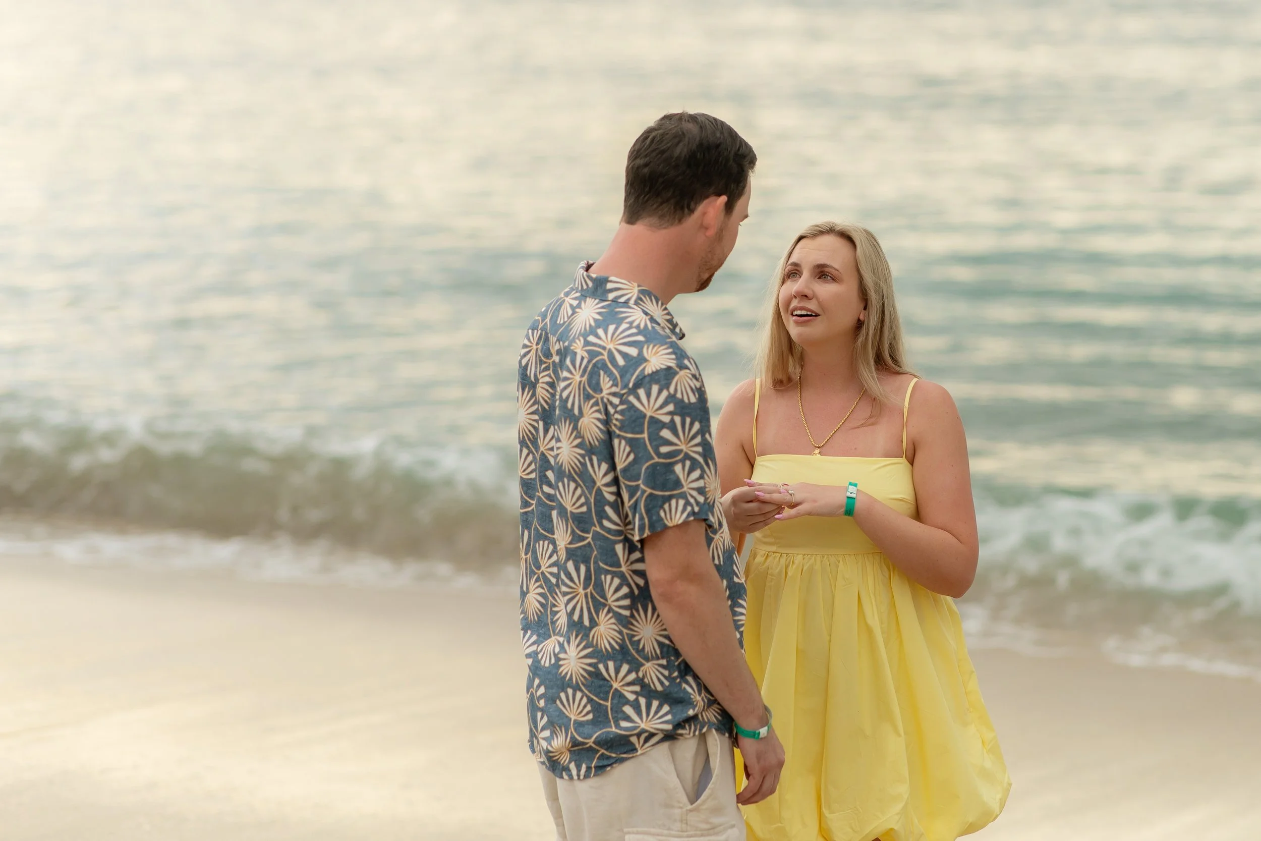 A couple having a heartfelt conversation on the beach with ocean waves in the background. The woman wears a yellow dress and the man wears a Hawaiian shirt. They are looking at each other emotionally.