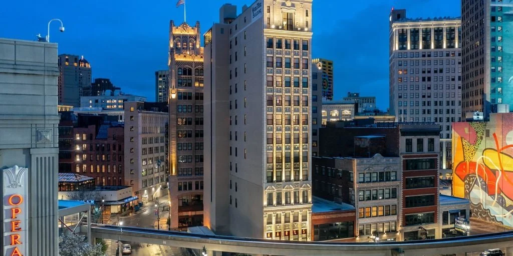 A cityscape with tall buildings illuminated at dusk, including a historic-looking building with a flag on top, and a colorful mural on a lower building.