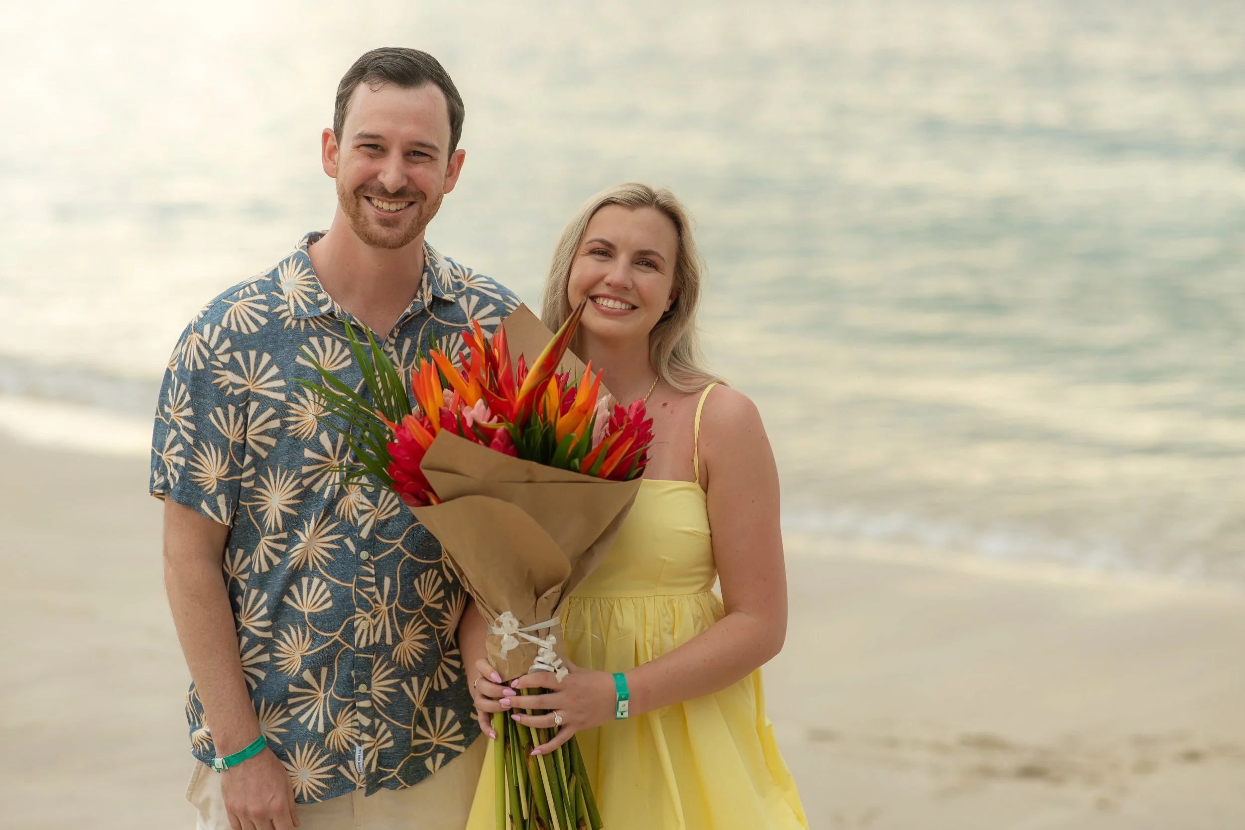 A smiling couple standing on a beach, holding a bouquet of colorful flowers, with the ocean in the background.