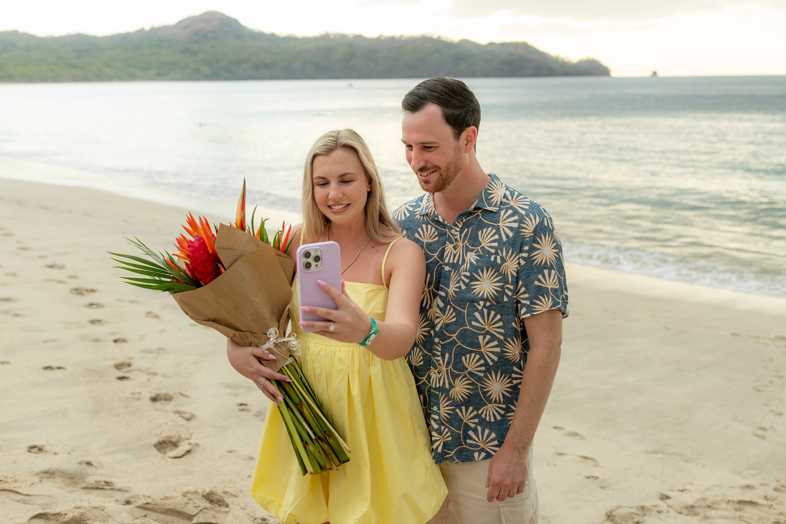 A woman in a yellow dress holding a bouquet of flowers, taking a selfie with a man in a Hawaiian shirt on the beach at sunset.