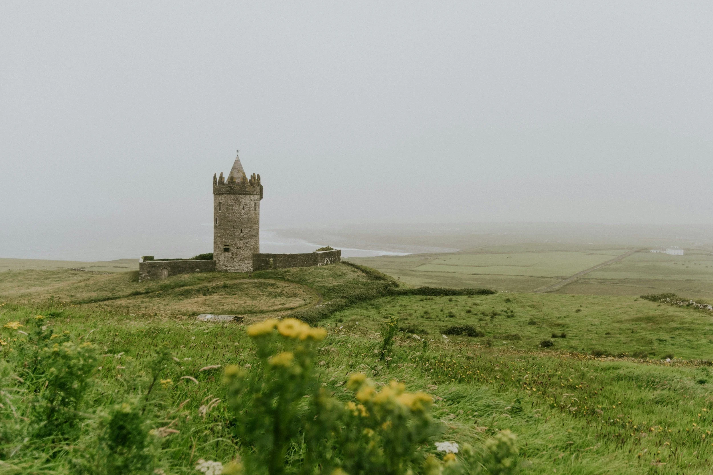 A stone tower on a grassy hill with a foggy overcast sky and fields in the background.
